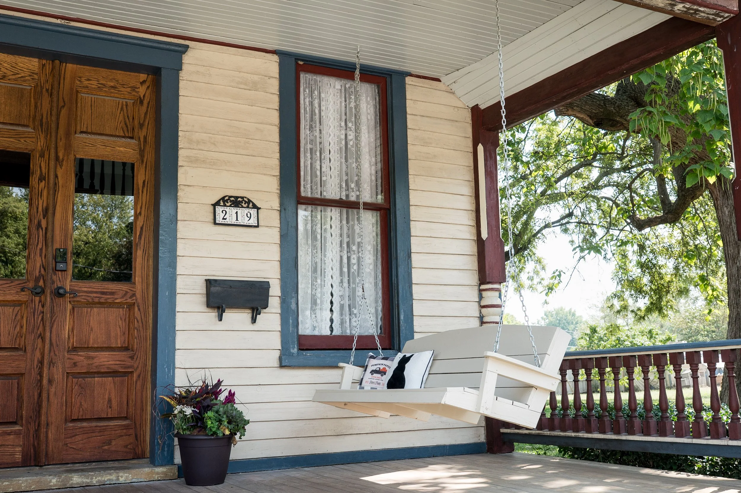 A porch with a wooden door, a window with lace curtains, a hanging swing, a potted plant, and a tree with green leaves in the background.