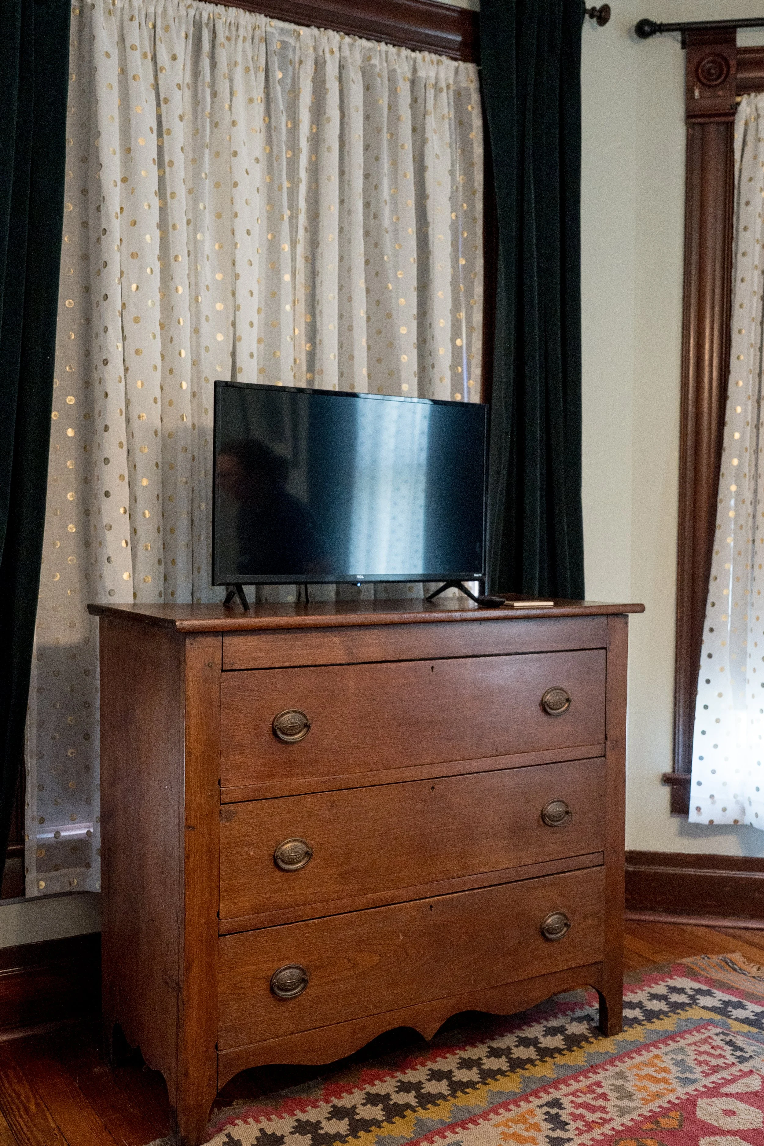 A tall wooden dresser with five drawers, each with oval-shaped metal handles, situated in a room with curtains featuring gold polka dots, a dark wooden trim, and an area rug with a colorful geometric pattern.