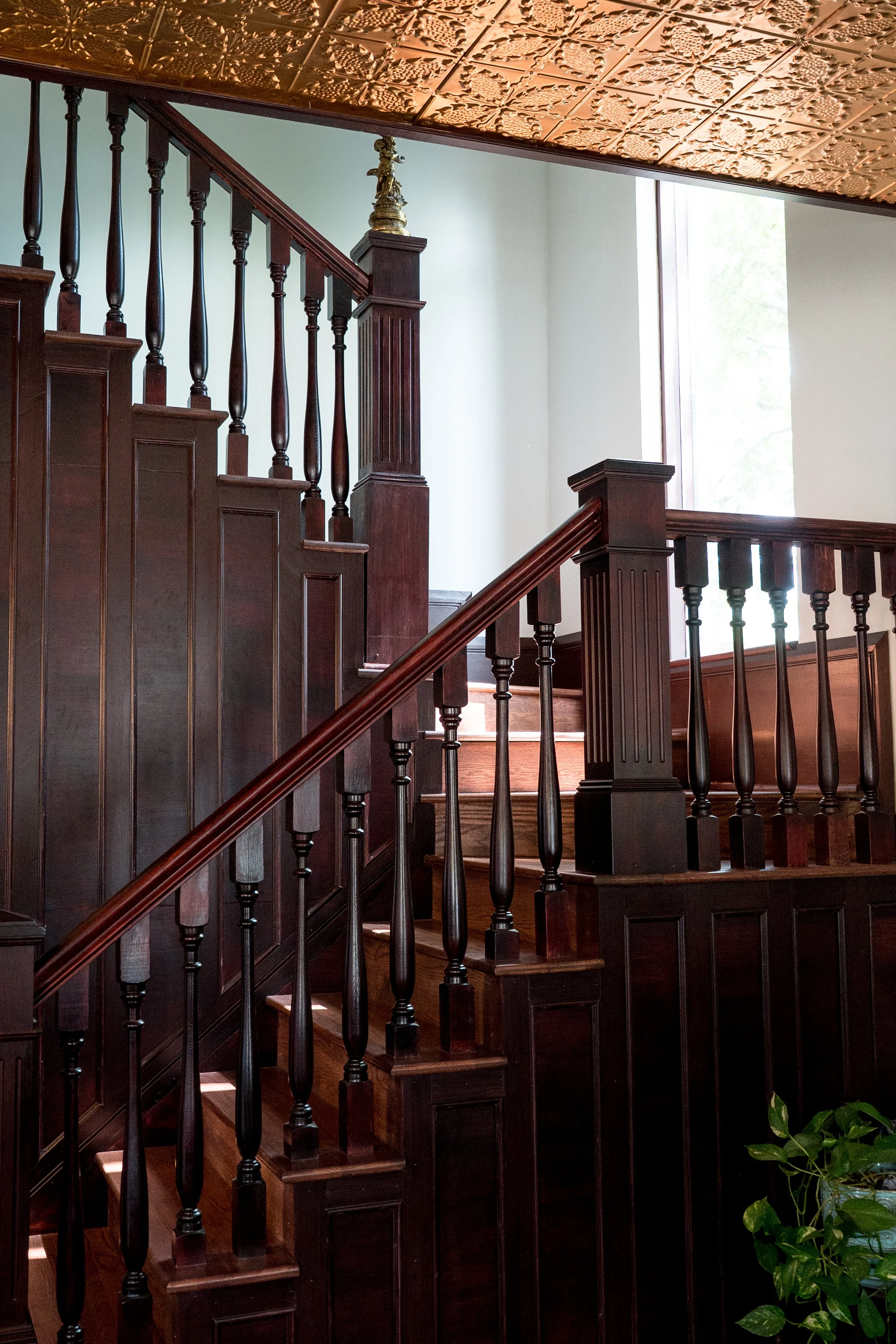 A wooden staircase with dark polished handrails and spindles, leading upward in a well-lit room by a large window, with a decorative ceiling pattern and a potted plant at the bottom right.