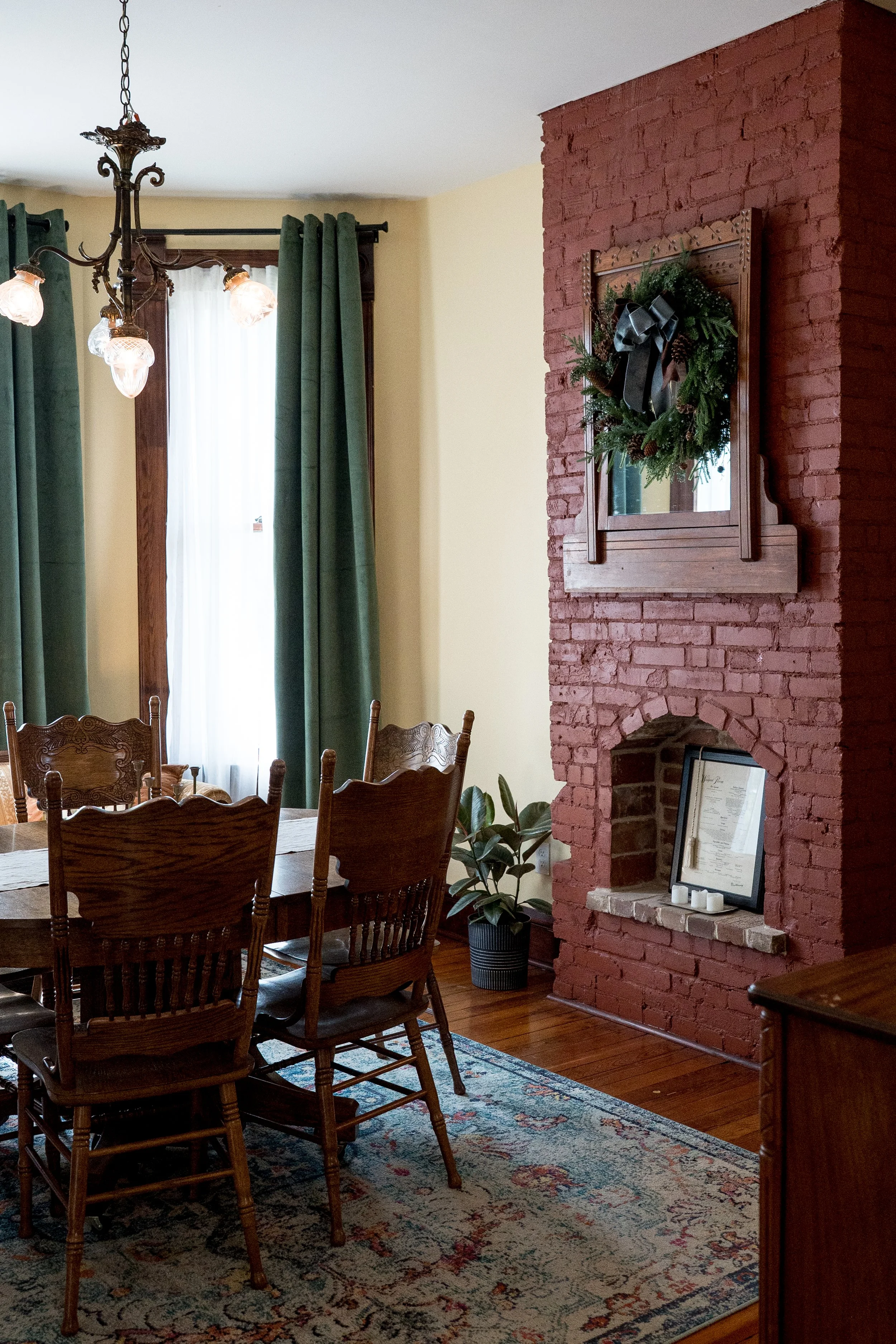 Dining room with wooden table and chairs, green curtains, a brick fireplace with a Christmas wreath, framed certificates, potted plant, hanging chandelier, and a patterned rug.