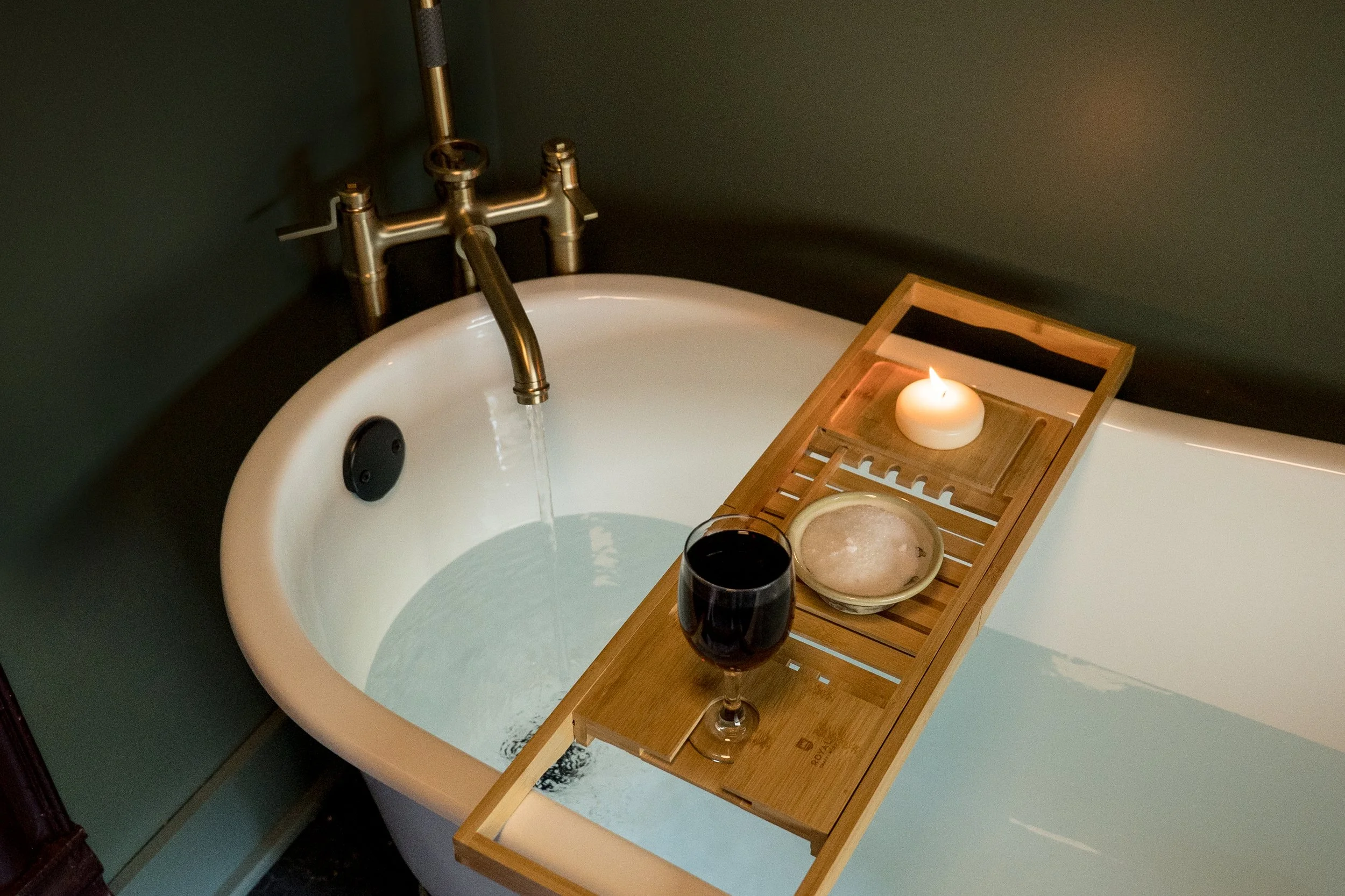 A bathroom sink with a brass faucet and hot and cold water knobs, a teal wall, a wooden bath tray holding a candle, a glass of red wine, and a dish with round soap.
