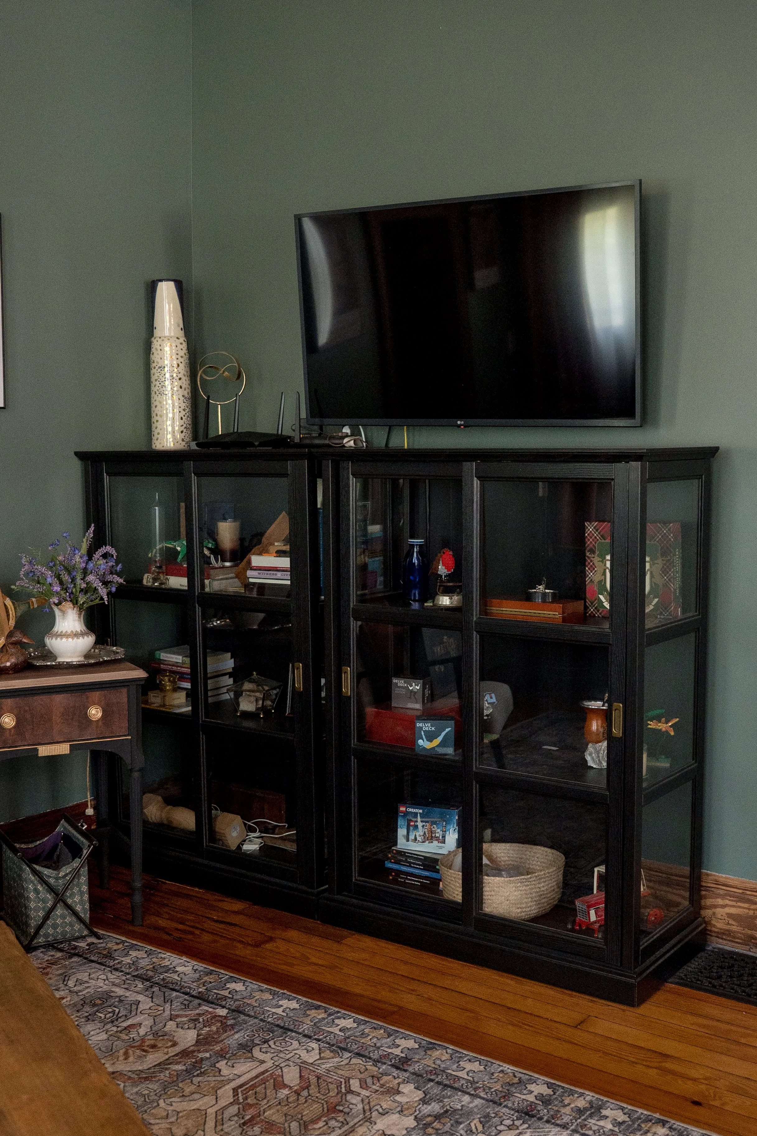 A black display cabinet with glass doors in a room with hardwood flooring. A large flat-screen TV is mounted above the cabinet. The room has a green wall, a patterned area rug, and a vase with purple flowers on a nearby table.