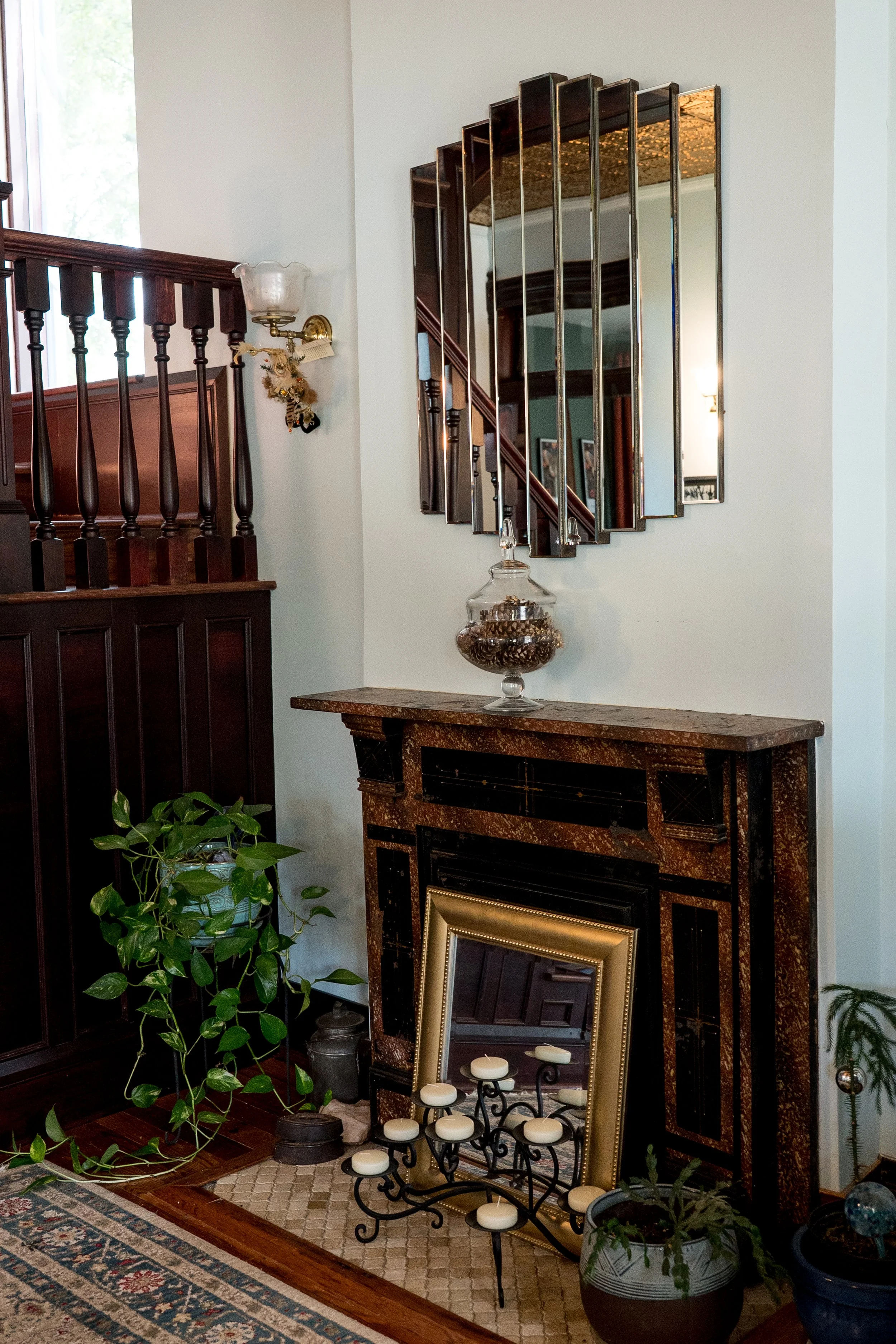 Interior of a living room with a wooden staircase, a decorative mirror on the wall, a fireplace with framed artwork above, and various potted plants.