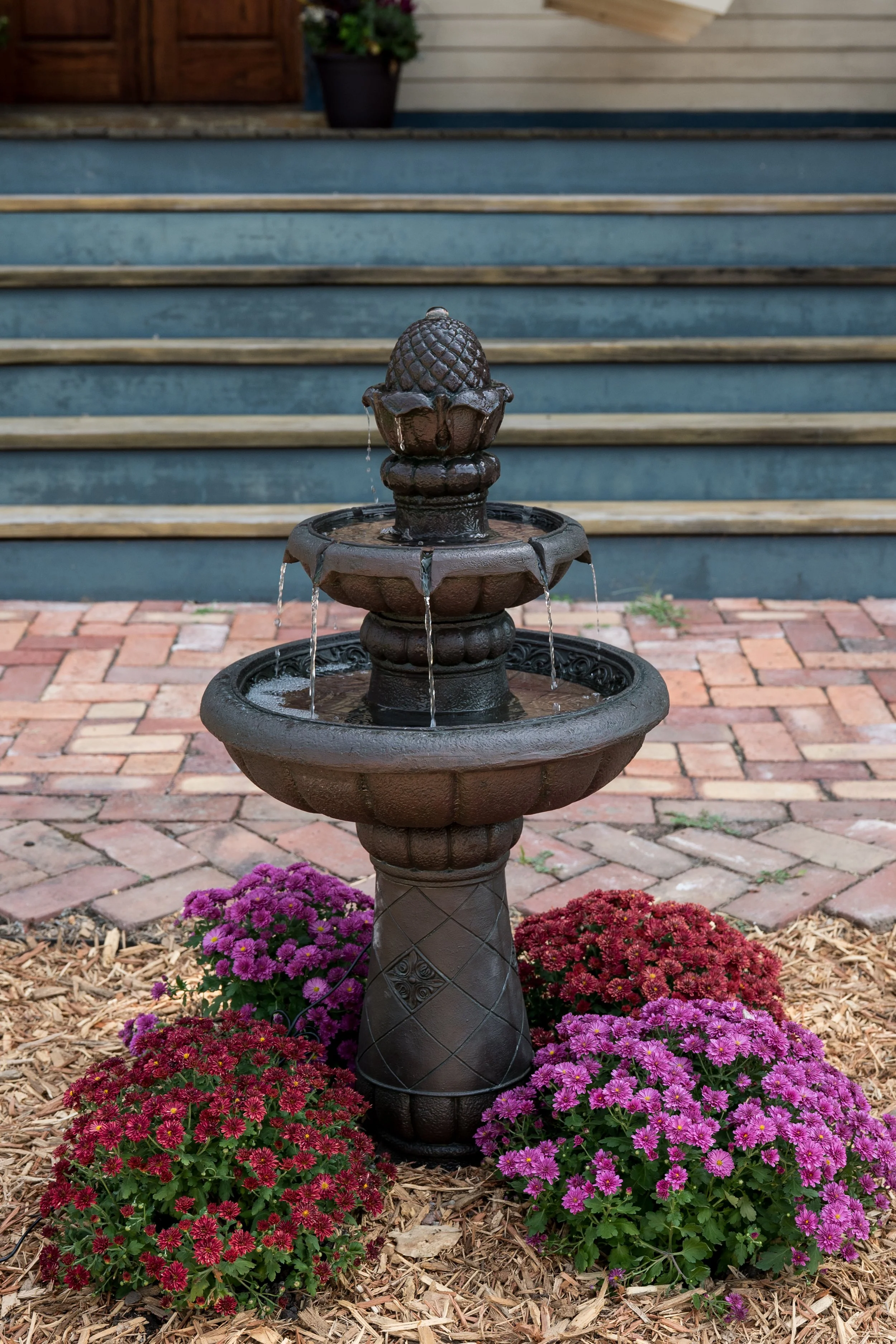 A decorative outdoor fountain made of dark metal, surrounded by pink, purple, and red flowers on mulch, with brick pavers and stairs with wooden railings in the background.