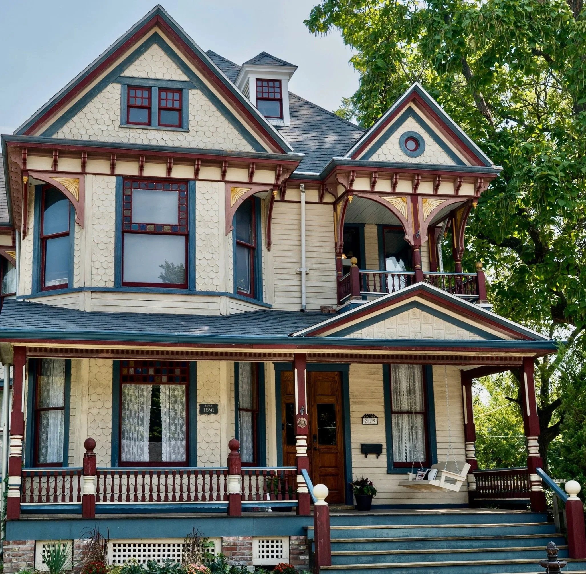 Victorian-style house with decorative trim, painted in cream, blue, and red, with a front porch, large windows, and a gabled roof, surrounded by greenery and trees.