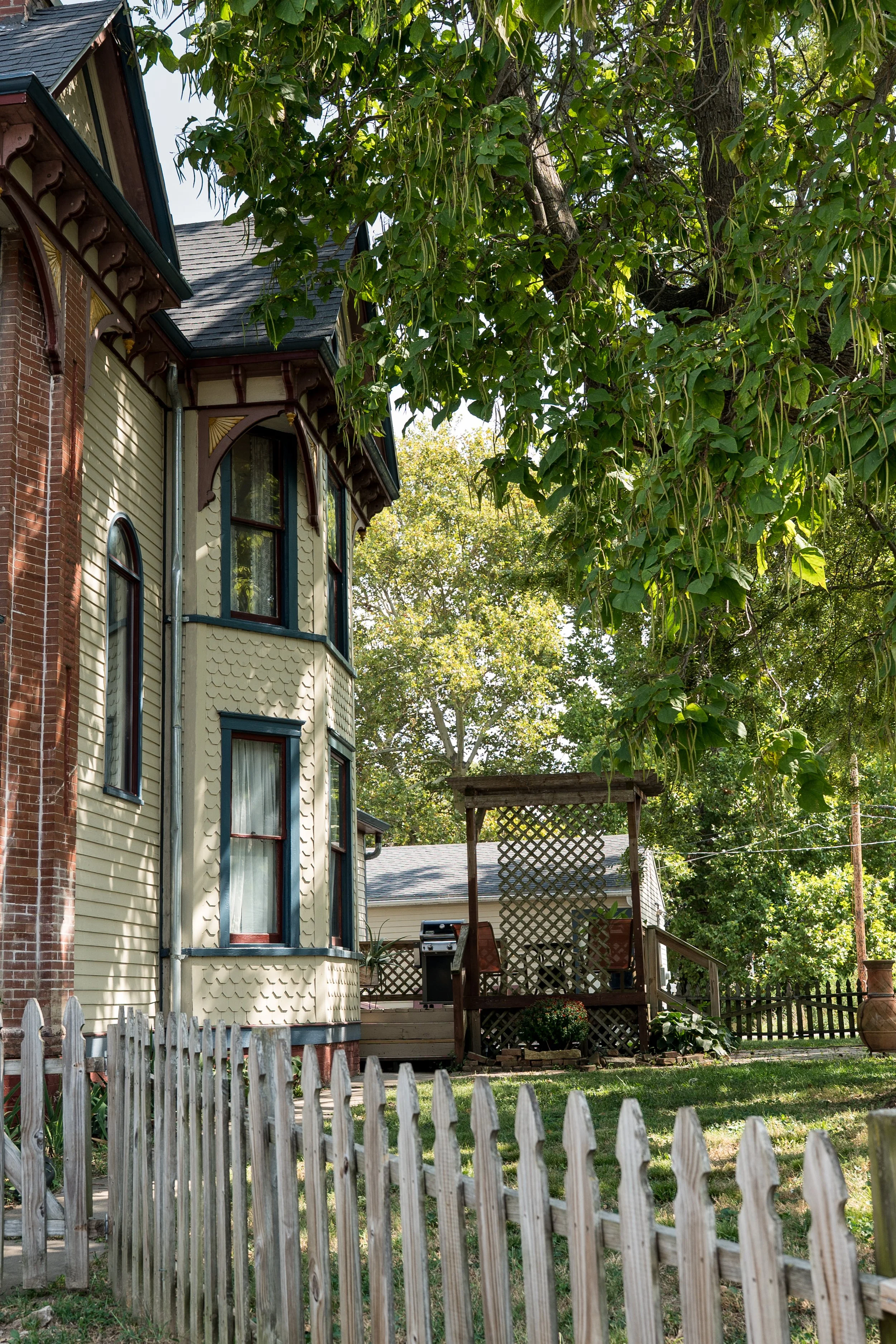 A Victorian-style house with beige siding, red brick accents, and dark trim, surrounded by a backyard with a white picket fence, lush green trees, and a small deck with lattice and outdoor furniture.