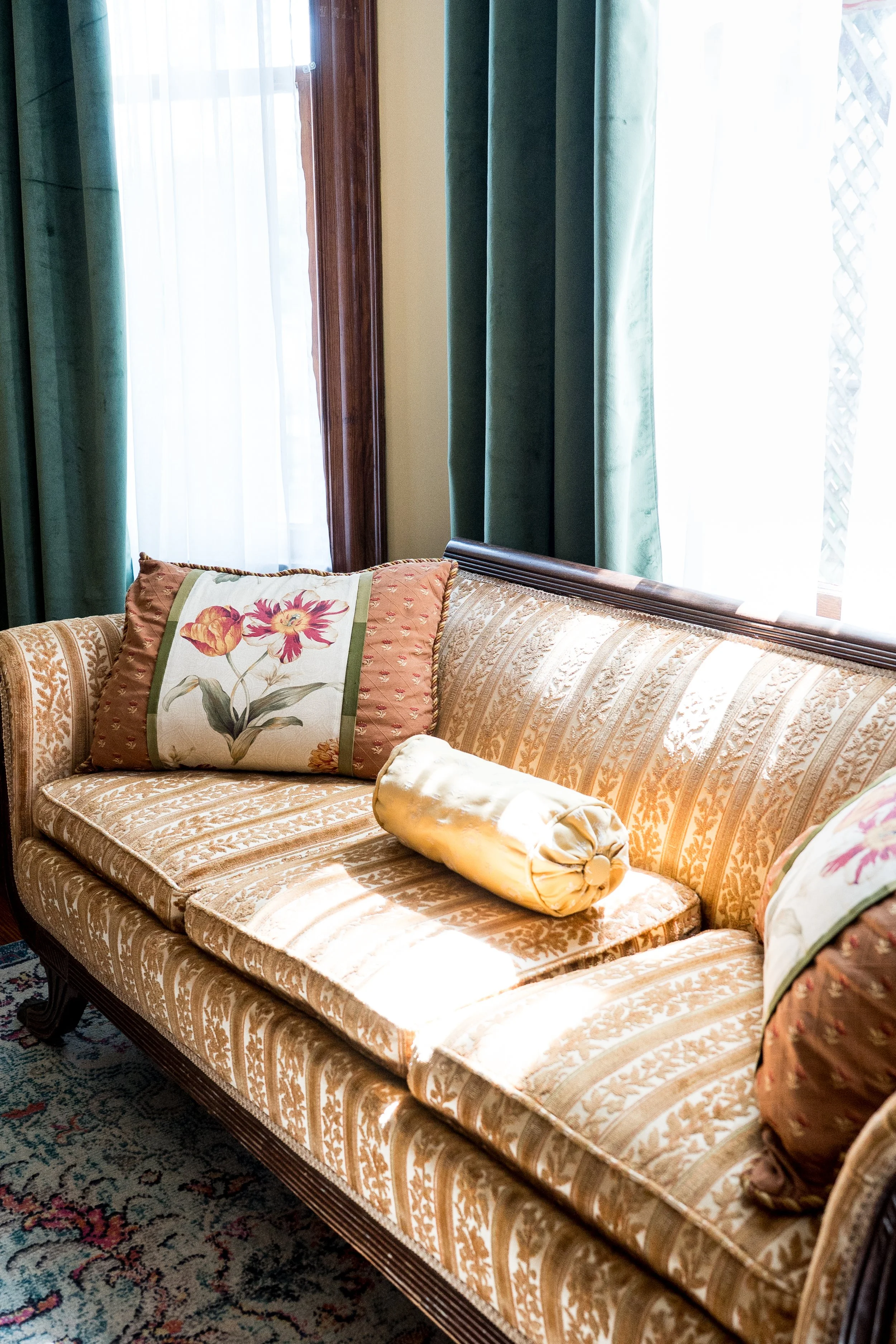 A vintage sofa with gold and cream patterned upholstery, a floral pillow, and a cylindrical bolster pillow, set against a background of window with green curtains and sheer white drapes, and a wooden window frame.
