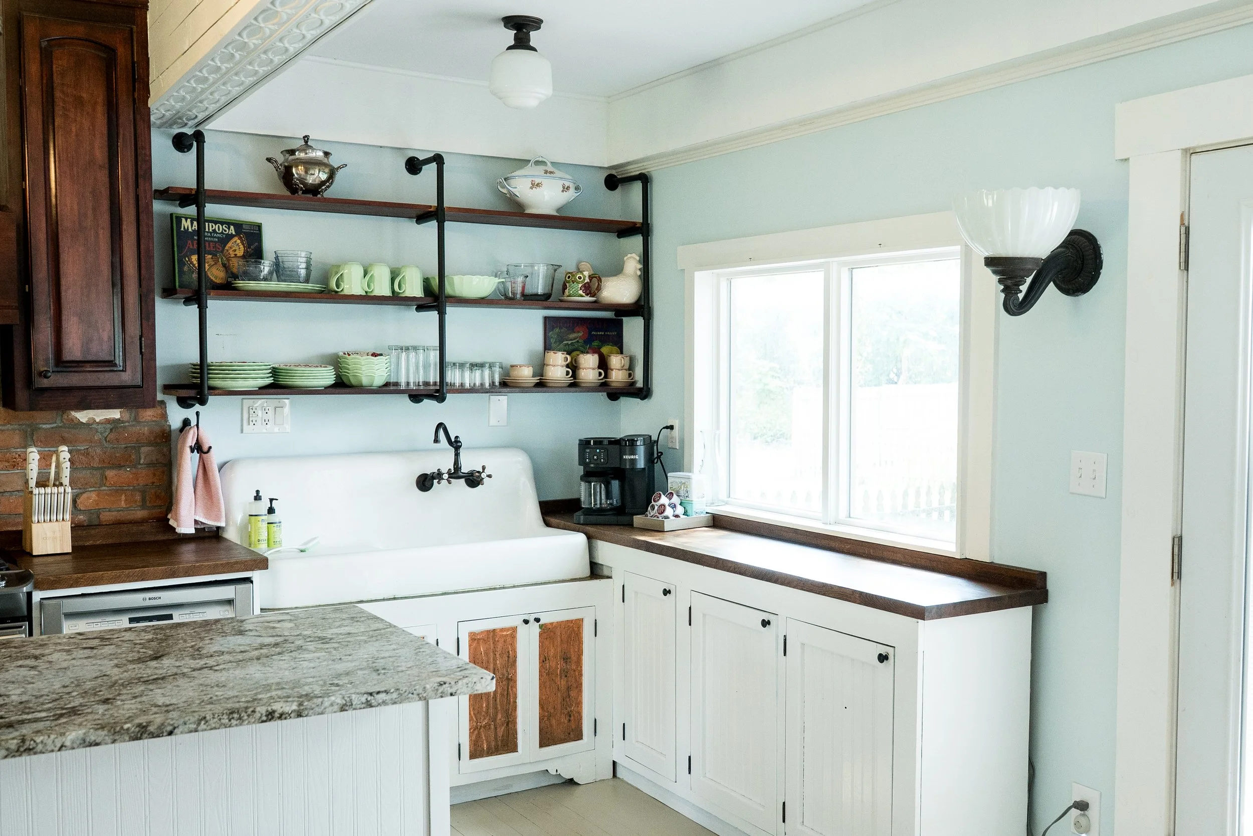 A cozy kitchen corner with open wooden shelves holding dishes, cups, and bowls, a utility sink with a vintage tap, and a coffee maker on the countertop by a large window, with a wall sconce light and a granite kitchen island in the foreground.