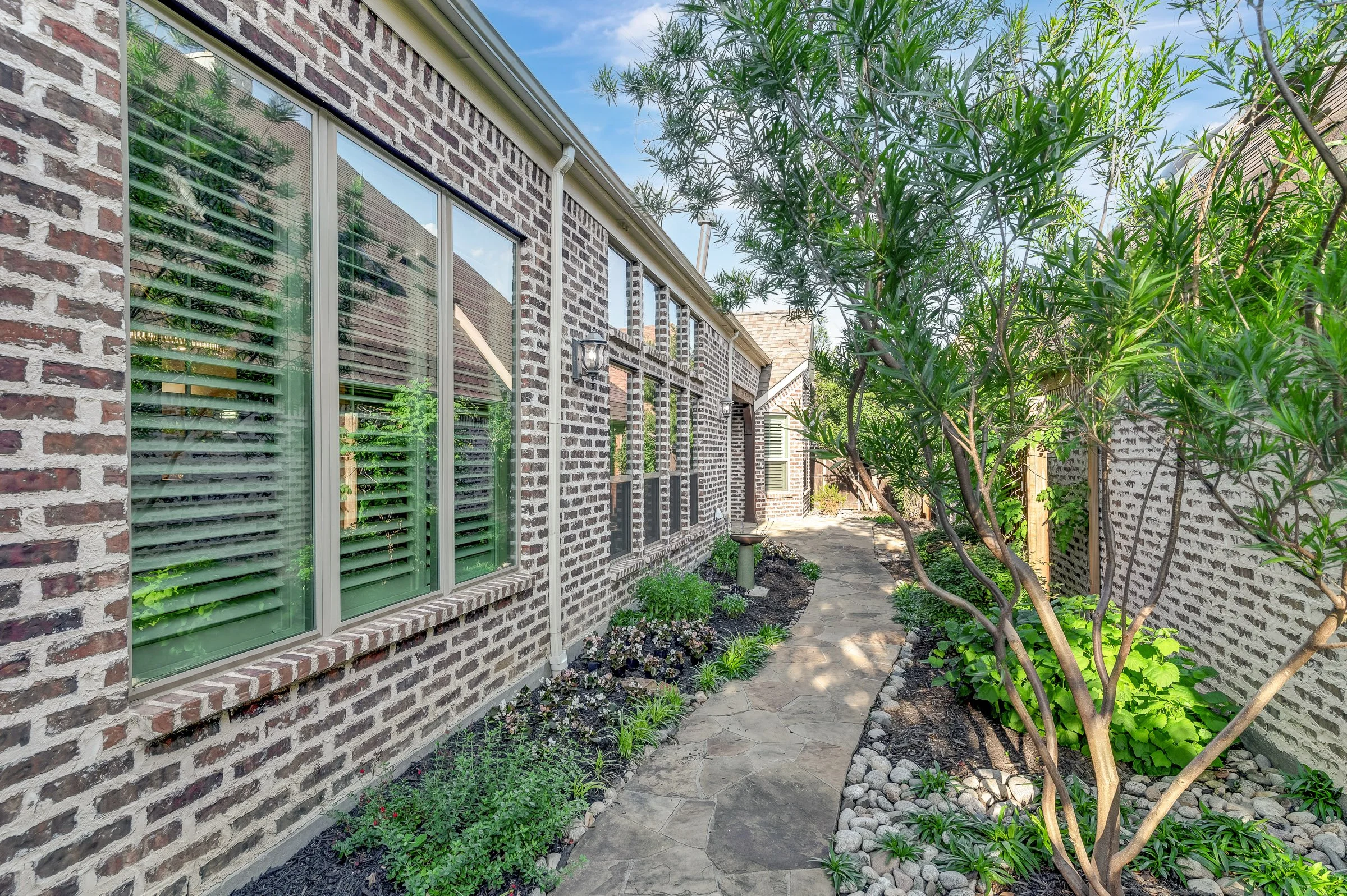 A brick house with large windows and green shutters on the left, with a garden path and lush greenery including trees and plants surrounding it.