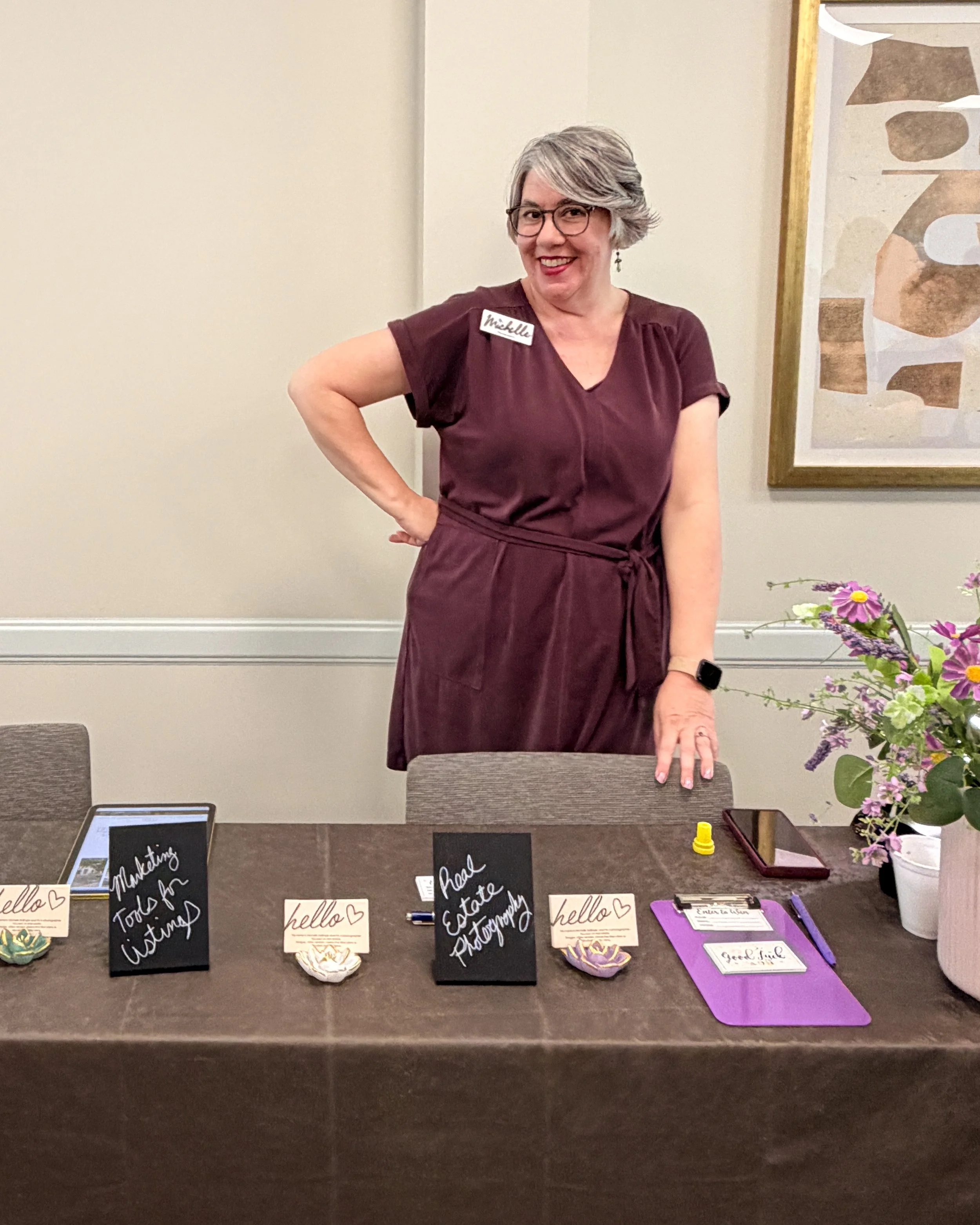 A woman with glasses and gray hair is standing behind a table at an event, smiling at the camera. She is wearing a maroon dress with a name tag that says "Michelle." The table has various signs and items, including a tablet, small chalkboards with handwritten messages, and a purple clipboard. There is a flower arrangement with pink and purple flowers on the right side of the table. In the background, there is a framed abstract artwork on the wall.