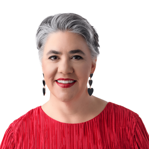 A woman with short gray hair, red lipstick, and black heart-shaped earrings, wearing a red top, smiling against a white background.