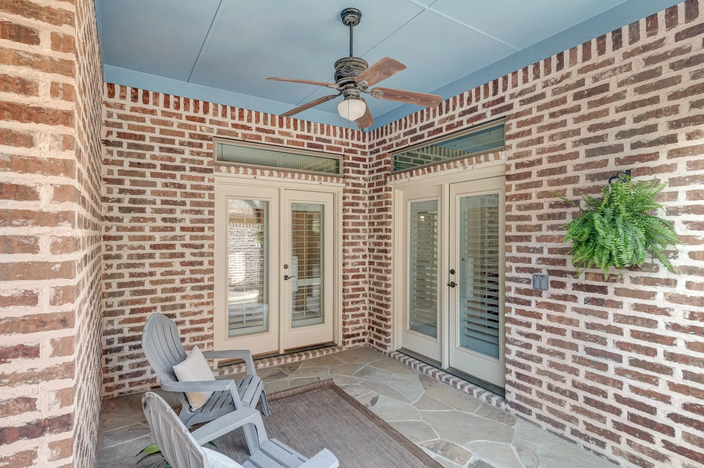A covered patio area with brick walls, a ceiling fan, glass double doors, a potted fern, two white chairs, and a rug on a stone floor.
