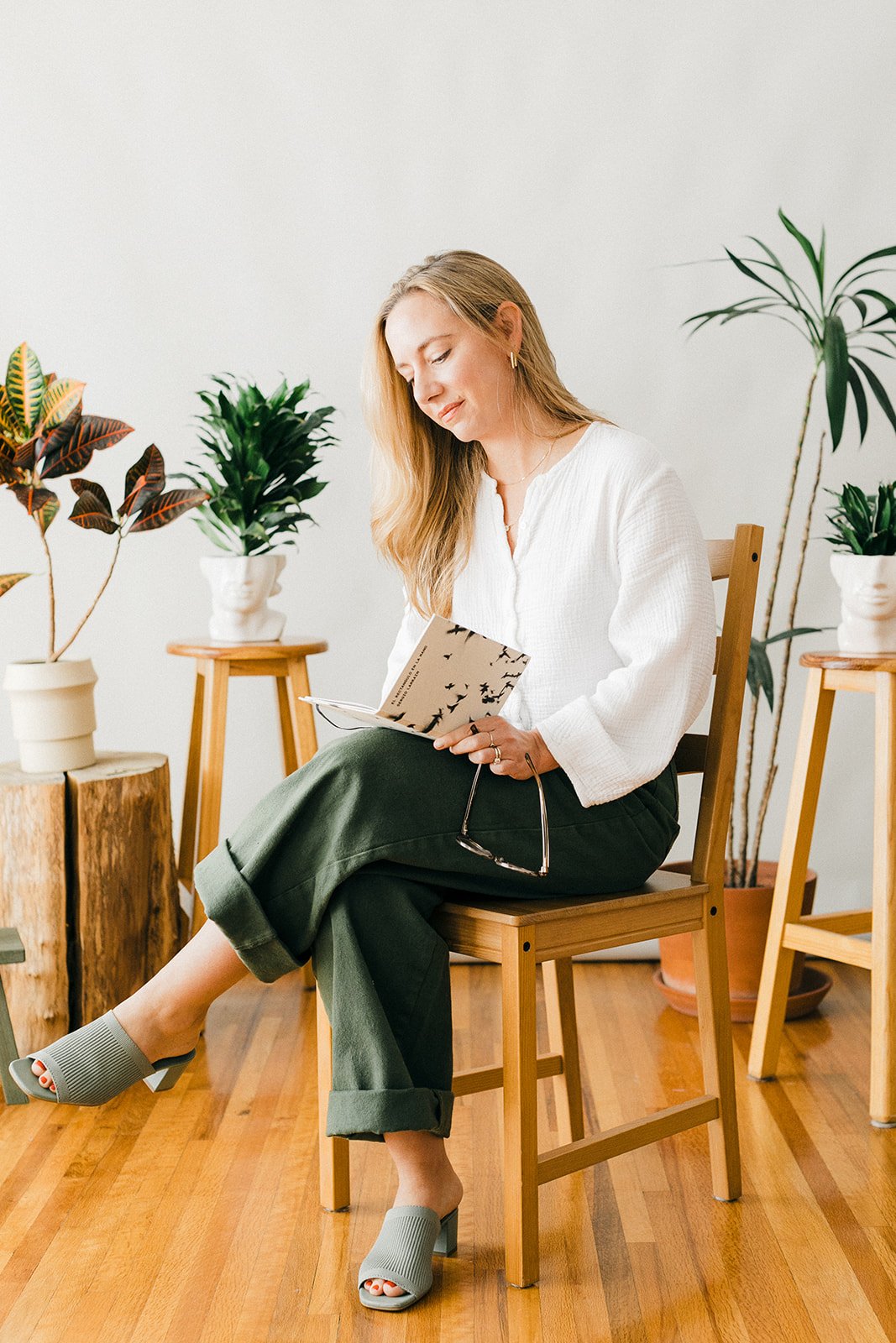 A woman sitting on a wooden chair, reading a book in a room with wooden flooring and several houseplants in pots around her.