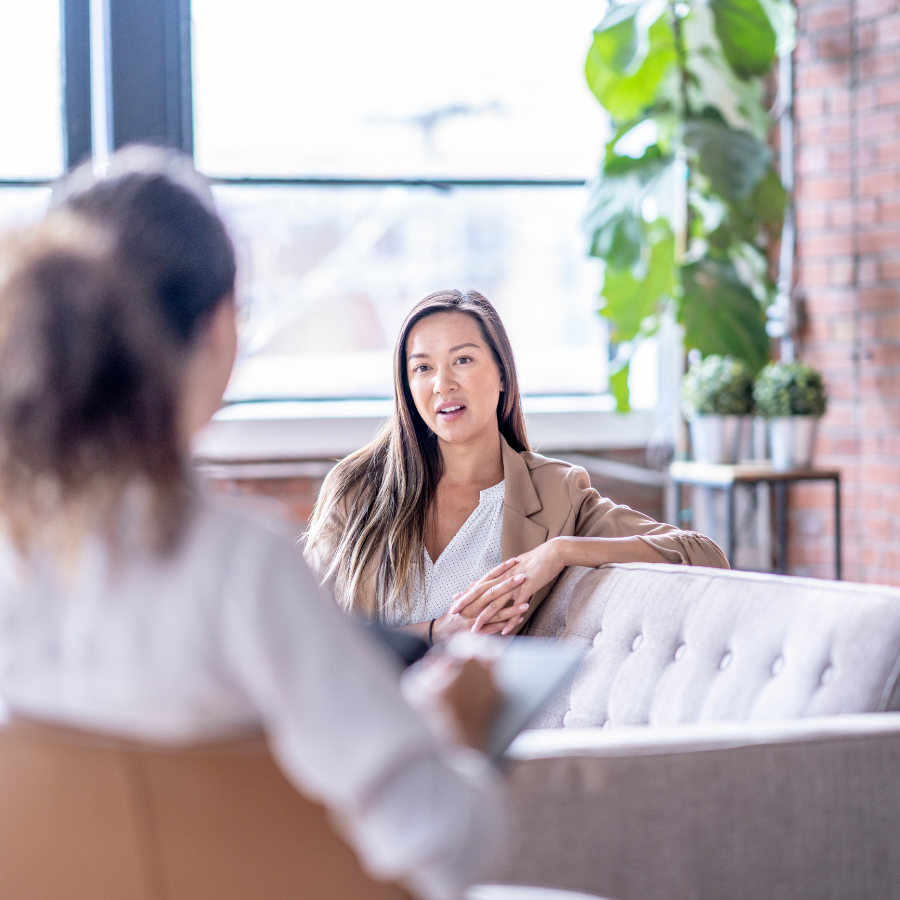 Two women having a discussion in a bright, modern office with large windows and indoor plants.