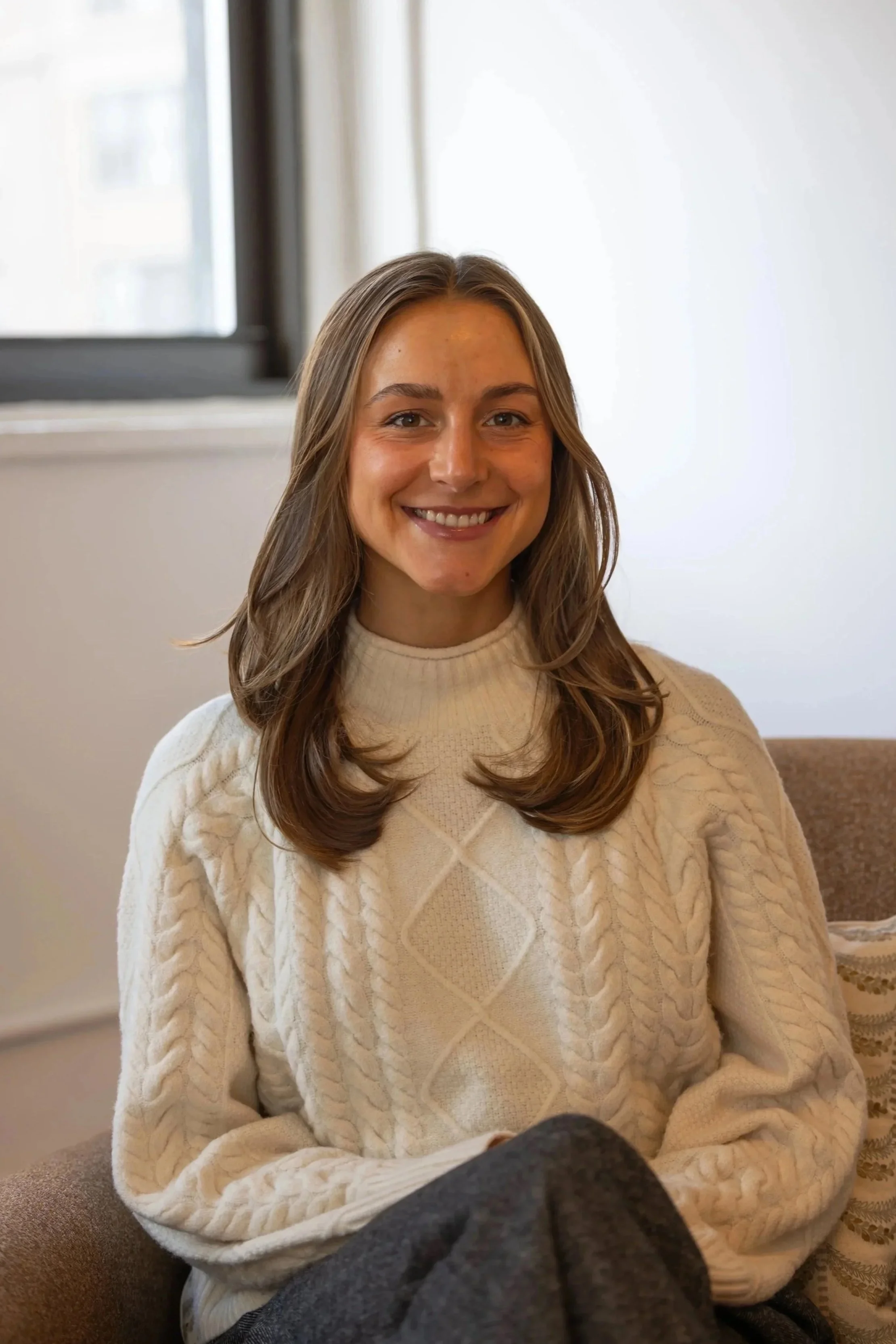A woman with long wavy hair smiling and wearing a cream-colored cable knit sweater, sitting indoors near a window with sheer curtains.