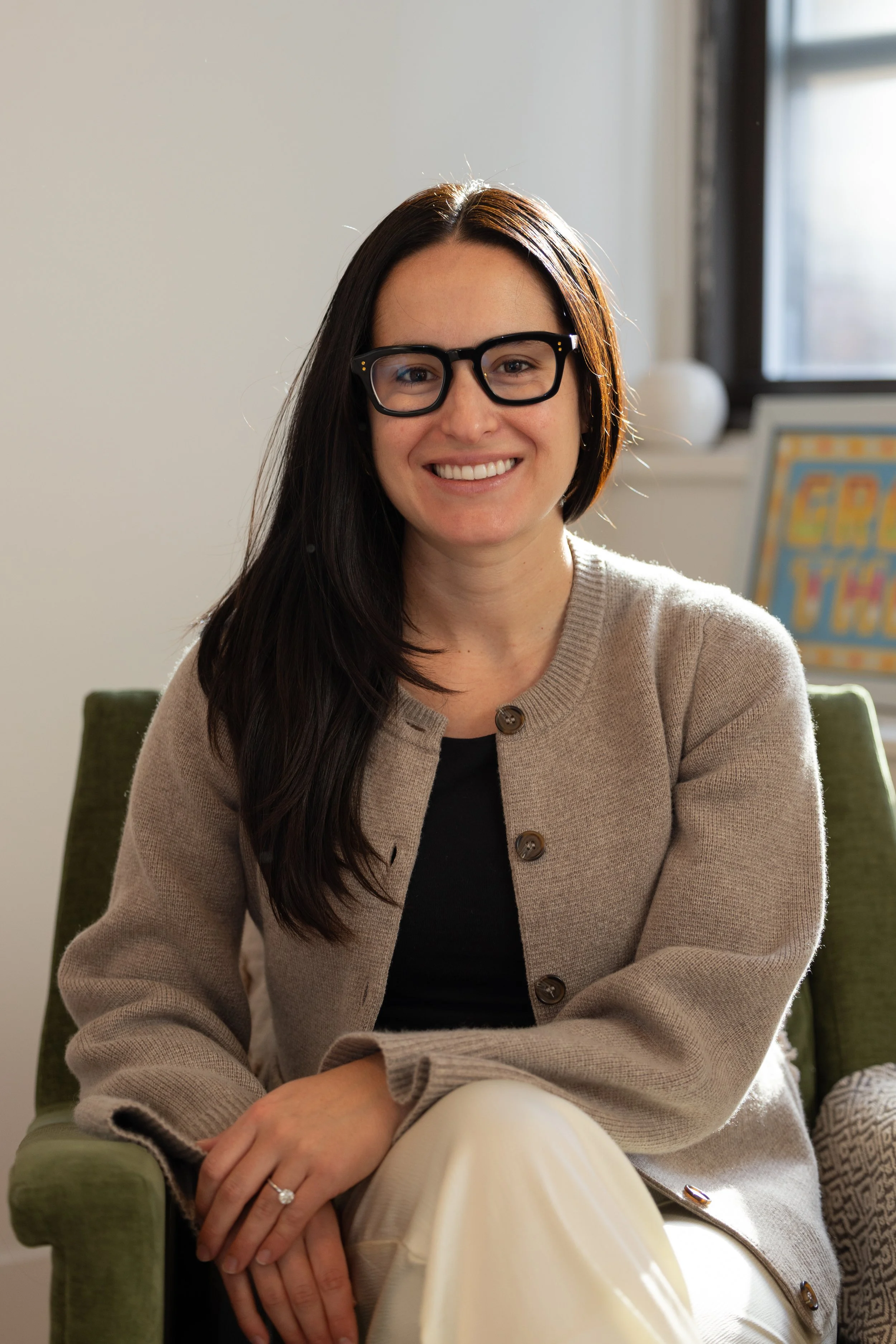A smiling woman with long dark hair, wearing glasses, a beige cardigan, and a black top, sitting on a green chair indoors near a window with natural light.