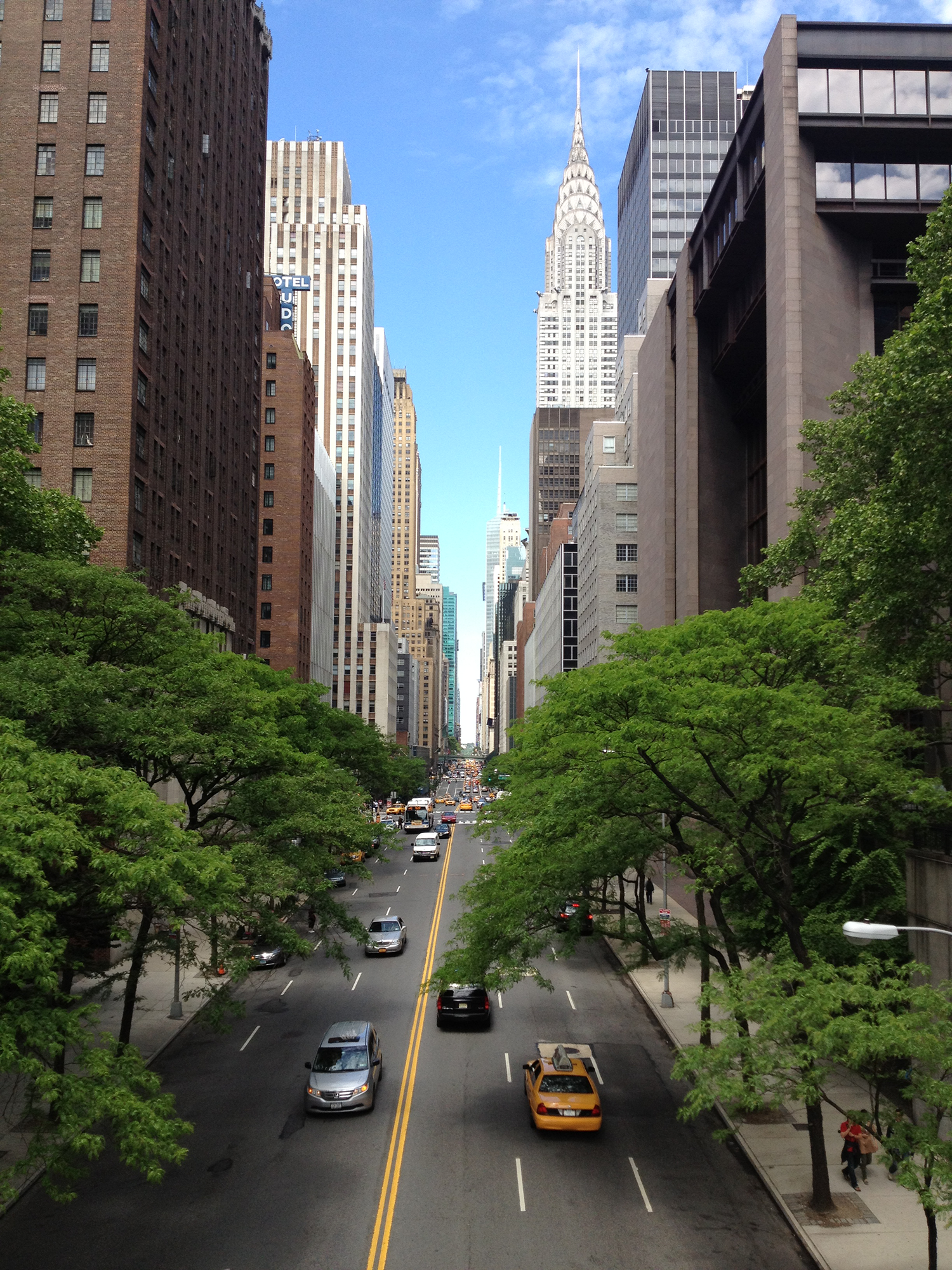 A city street flanked by tall buildings, with parked cars and moving vehicles, and green trees along the sidewalks. The sky is blue with some clouds.