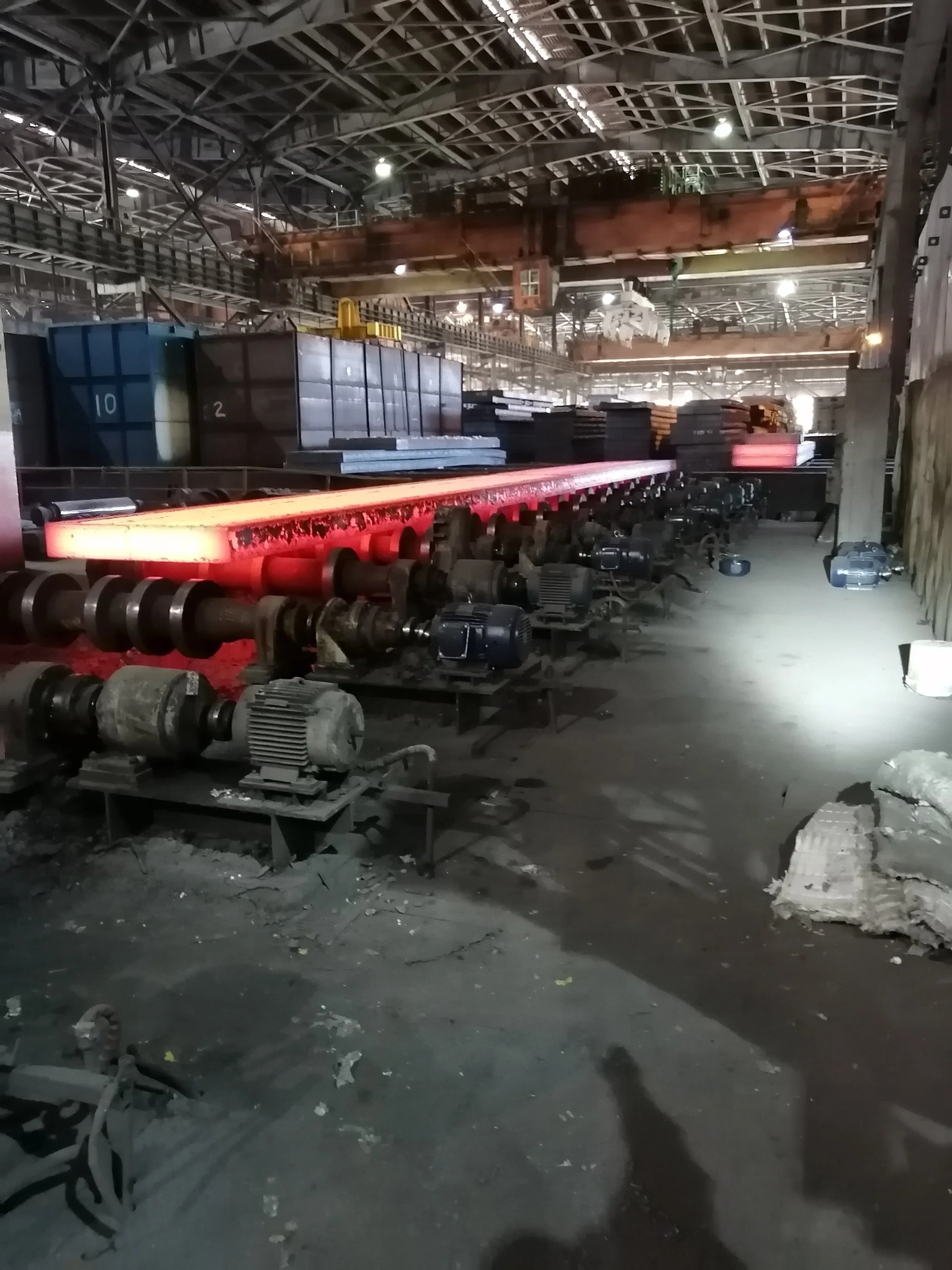 Interior de un taller industrial con maquinaria y equipos. Hay una línea de máquinas en primer plano y estructuras metálicas en el techo, con iluminación artificial y objetos dispersos en el suelo.