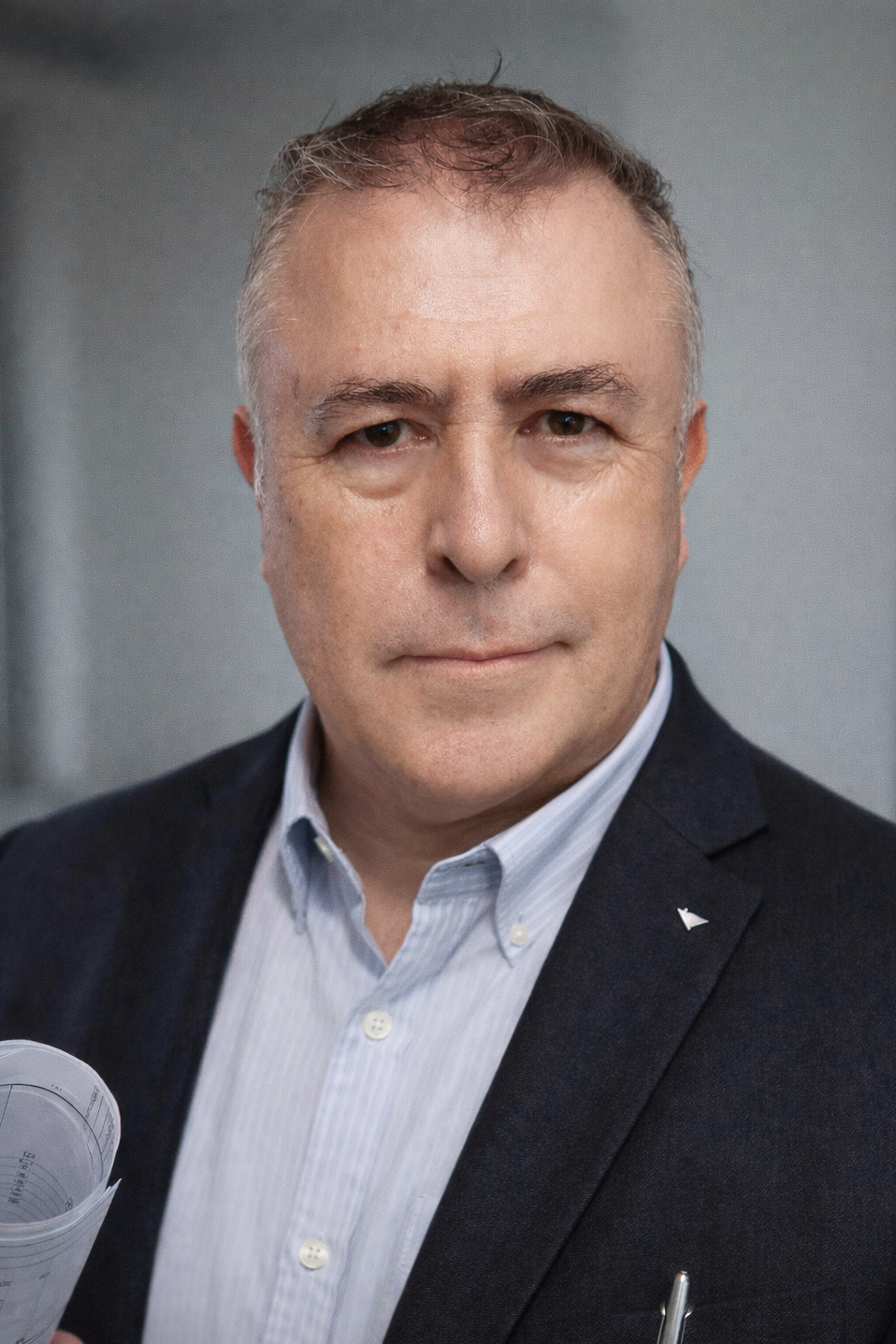 Hombre con cabello gris y expresión seria, usando saco negro y camisa blanca, sosteniendo unos papeles en una mano.