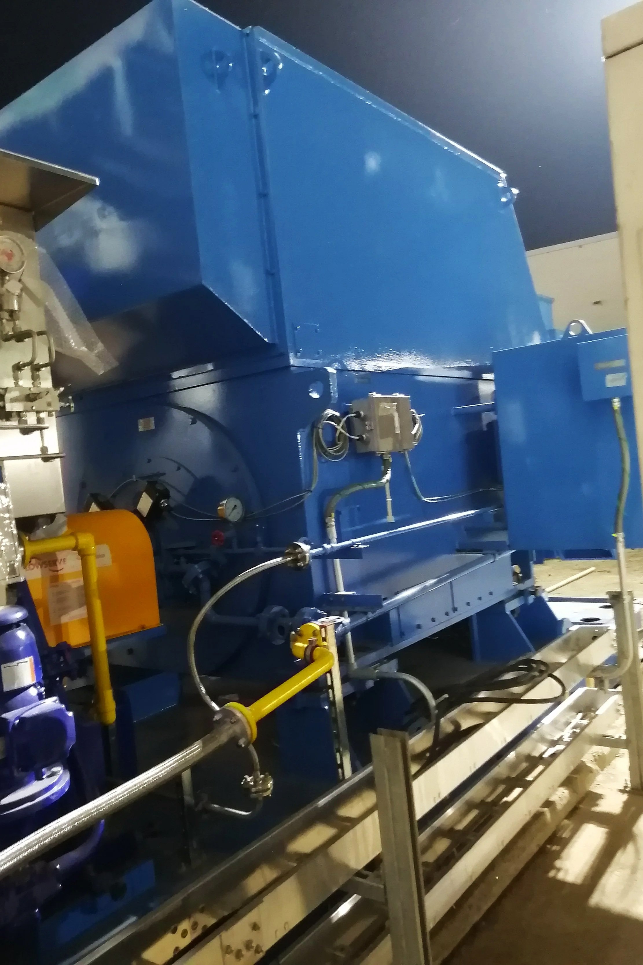 Imagen de una máquina industrial grande de color azul en un entorno de fábrica, con tuberías, válvulas y componentes metálicos.