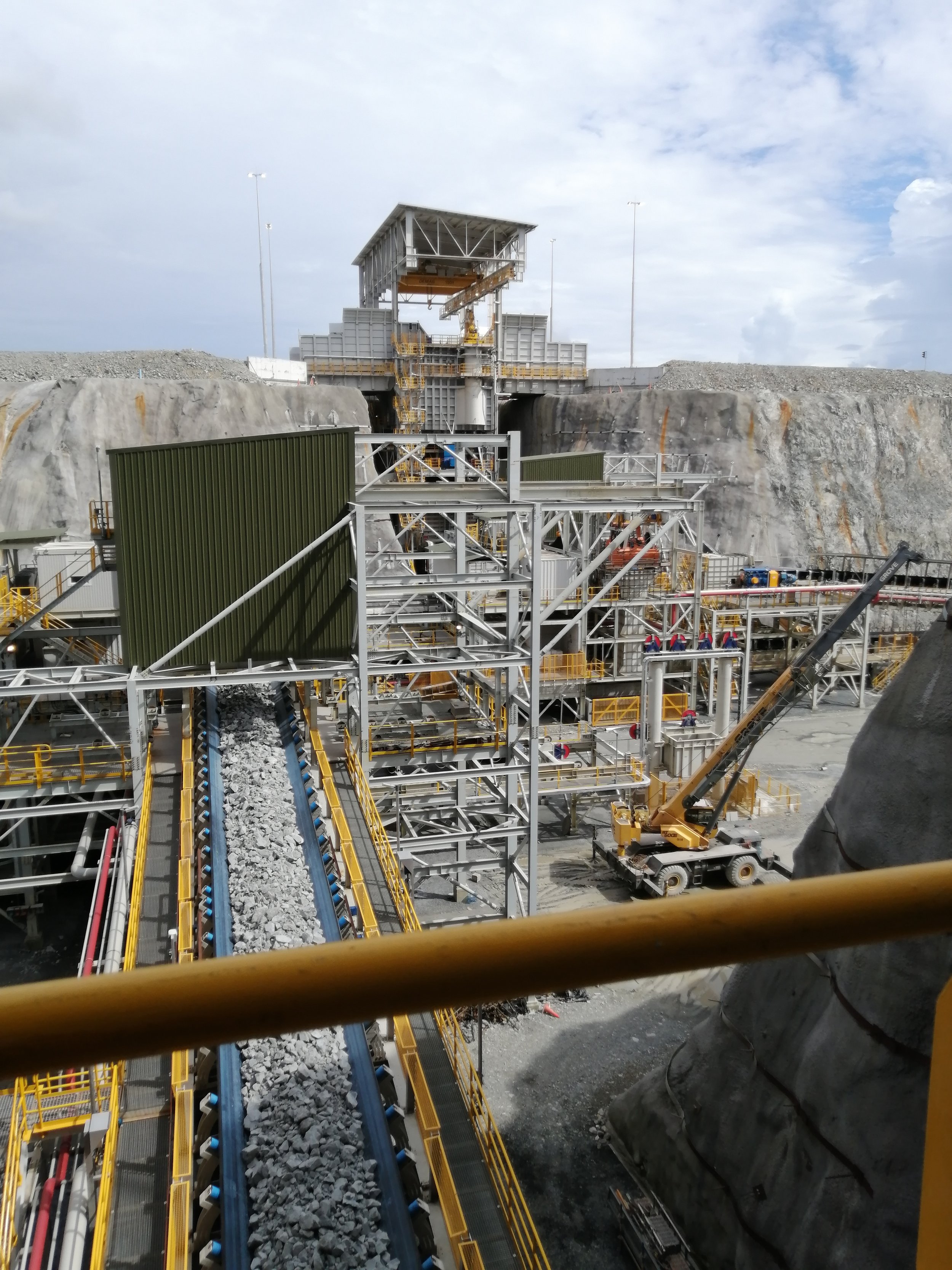 Una estructura de construcción industrial con maquinaria, cintas transportadoras y una montaña de piedra, en un entorno al aire libre con cielo parcialmente nublado.