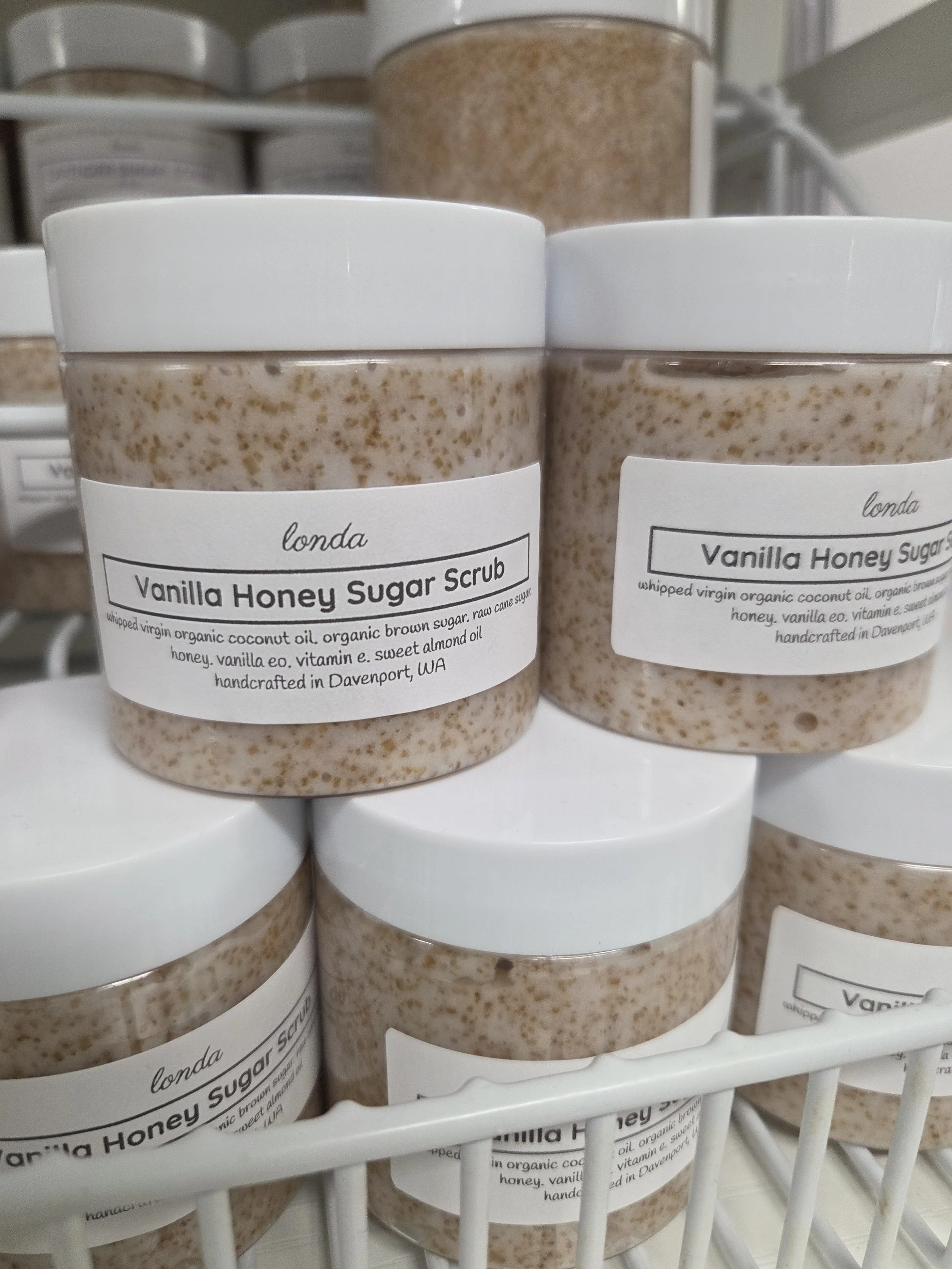 Close-up of containers labeled 'Vanilla Honey Sugar Scrub' with white lids, sitting in a white wire basket on a store shelf.