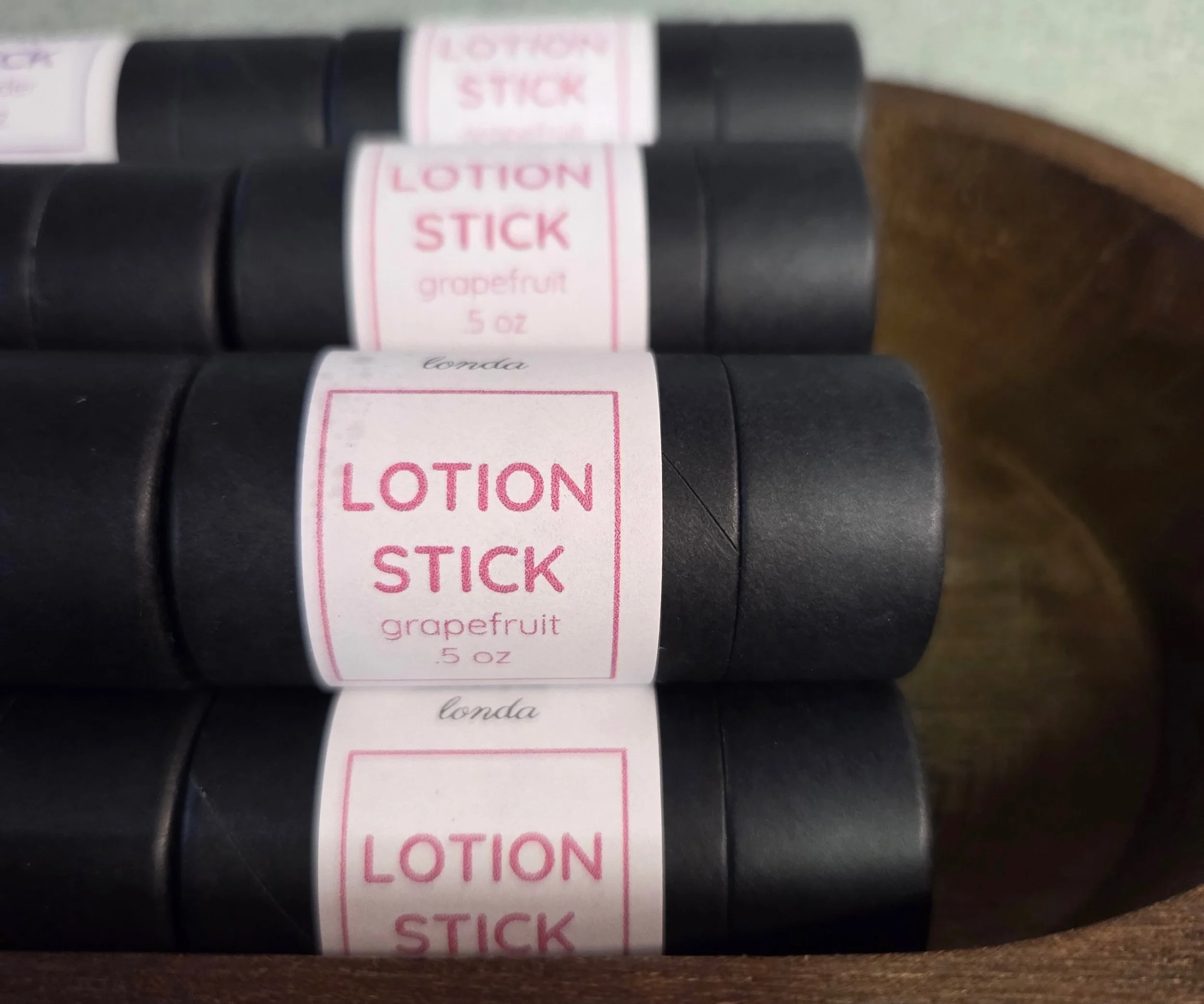 Three black lotion sticks with pink labels reading 'Lotion Stick, grapefruit, 0.5 oz,' placed in a wooden container.
