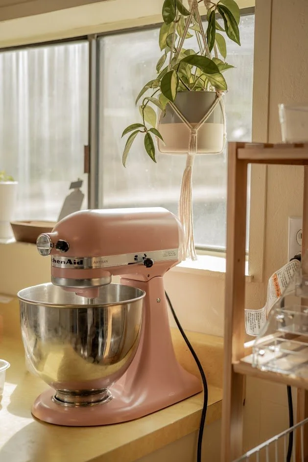 A pink KitchenAid stand mixer on a wooden countertop next to a window, with a hanging potted plant above it and a small wooden shelf on the right.