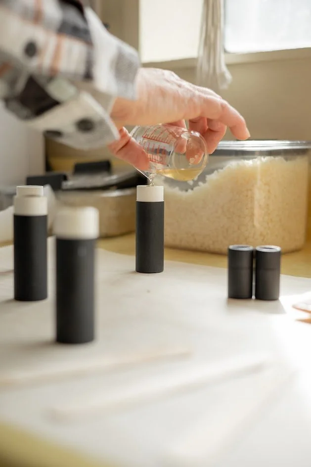 Close-up of a person pouring liquid from a small glass beaker into a test tube on a kitchen countertop, with several black-capped test tubes and a large bowl of rice in the background.