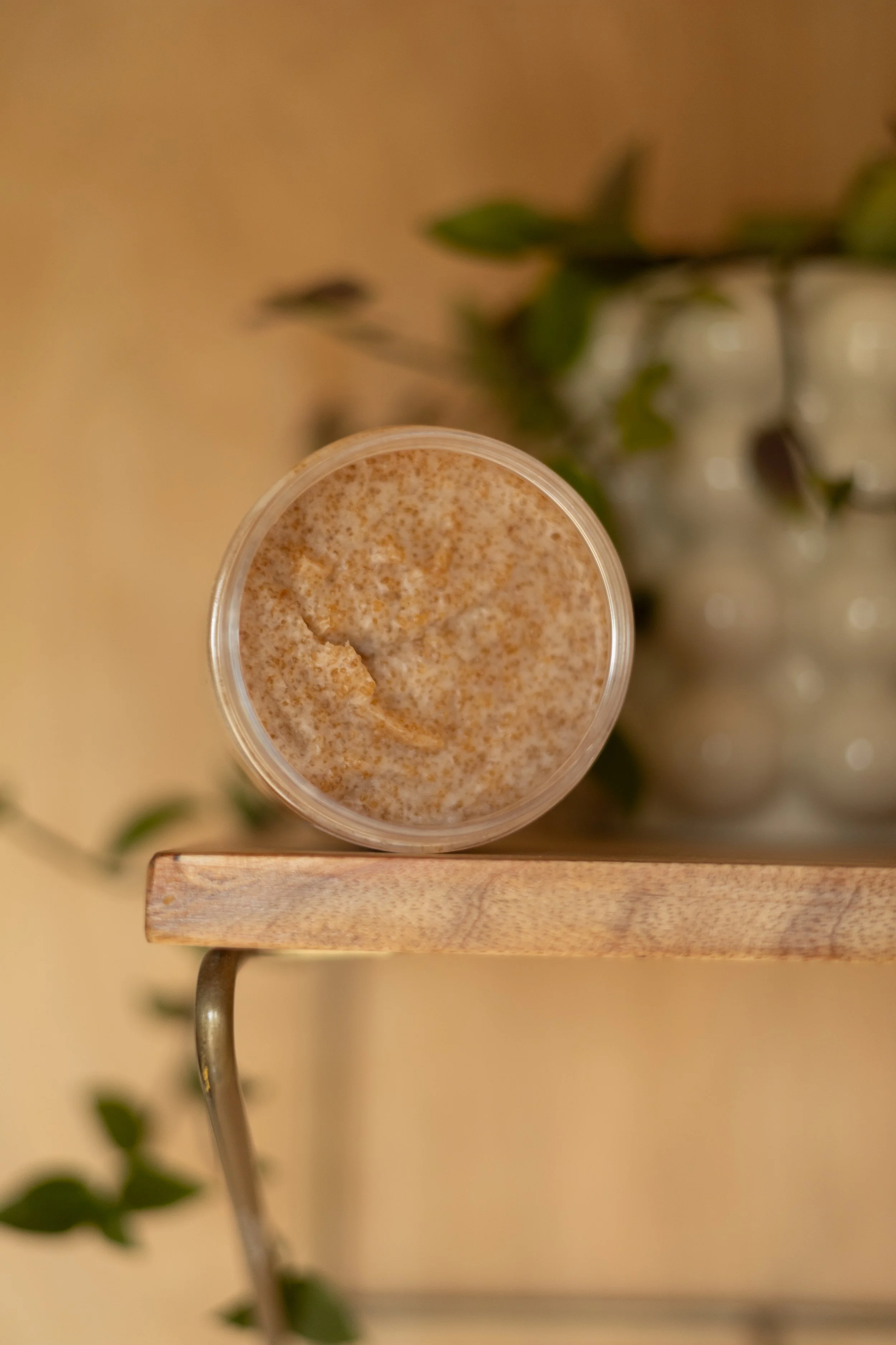 Top-down view of a glass filled with a frothy, creamy beverage, placed on a wooden surface with a blurred background of green leaves and a jar of white decorative balls.