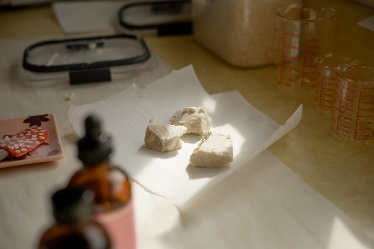Three pieces of white substance on a crumpled white paper on a kitchen counter, with measuring cups and small bottles nearby.