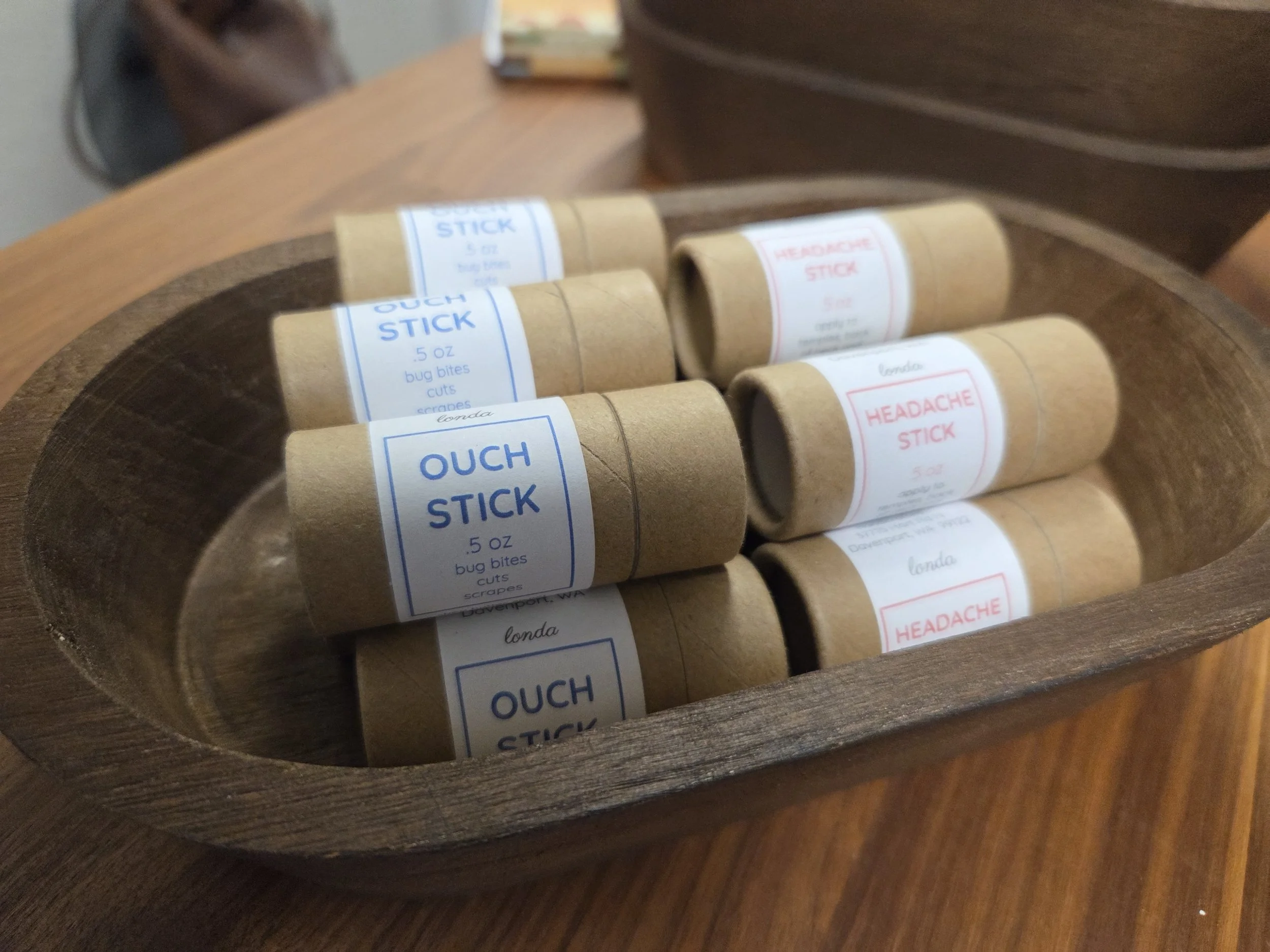 A wooden bowl containing six small cylindrical tubes labeled as headache sticks and ouch sticks, used for bug bites, cuts, and scrapes.