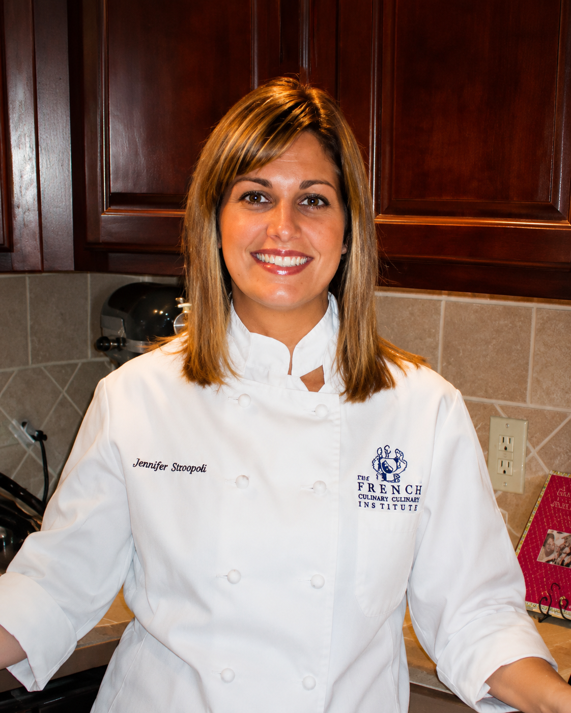 A woman in a white chef's coat standing in a kitchen, smiling at the camera