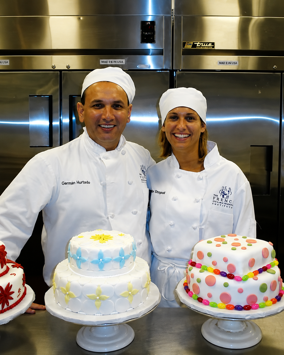 Two pastry chefs, a man and a woman, wearing white chef uniforms and hats, standing in front of a stainless steel kitchen. They are smiling and holding decorated cakes on display stands.
