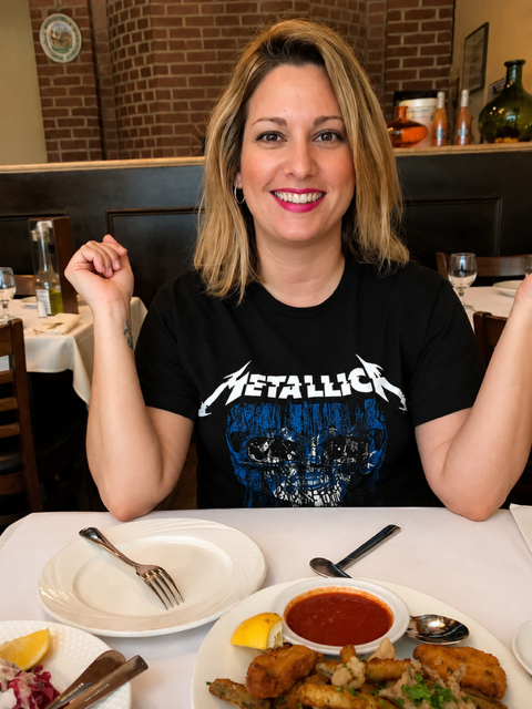 A woman with shoulder-length blonde hair and a black Metallica t-shirt sits at a dining table in a restaurant with brick walls, smiling at the camera. There are plates, utensils, and a bowl of sauce on the table.