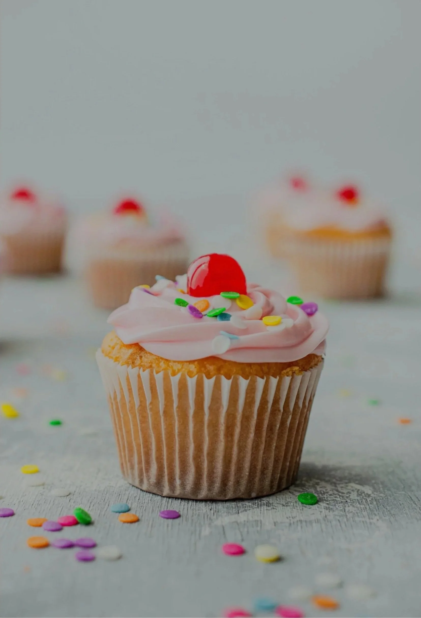 A cupcake with pink frosting, a cherry on top, and colorful sprinkles, placed on a gray surface with additional sprinkles scattered around. There are three blurred cupcakes in the background.