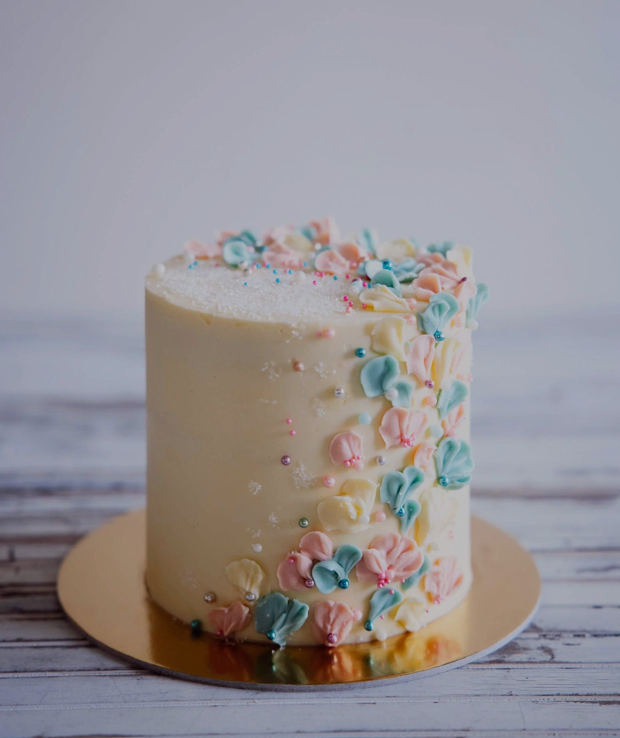 Close-up of a white frosted birthday cake decorated with pastel-colored icing flowers and small edible pearls, placed on a gold-colored cake board on a wooden surface.