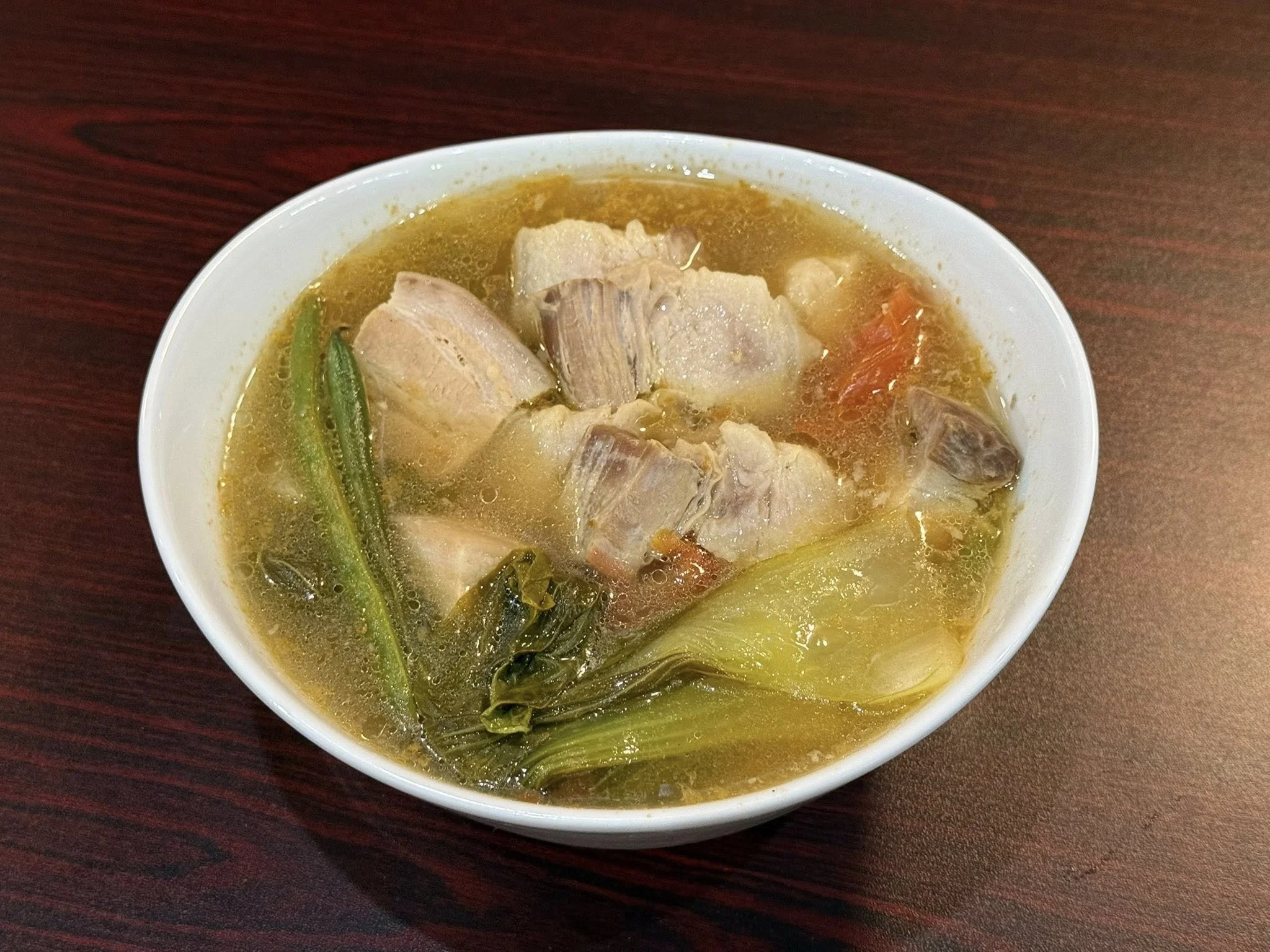A bowl of soup with chunks of pork, green vegetables, and a clear broth on a dark wooden table.
