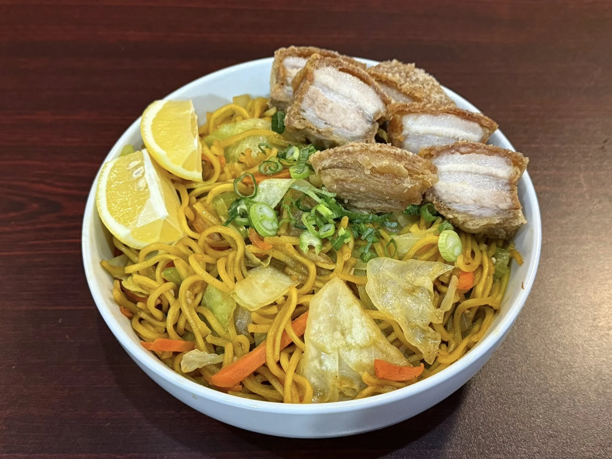 Bowl of fried noodles with vegetables, lemon wedges, and pieces of fried pork belly on top.