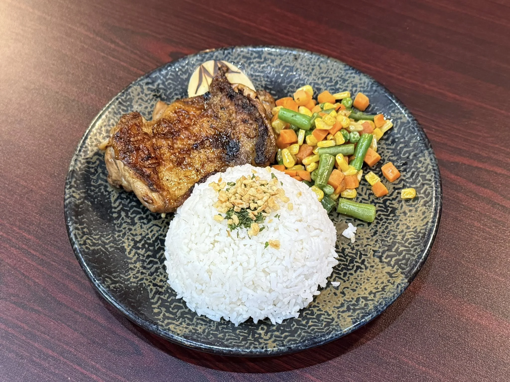 Plate with a piece of grilled chicken, a serving of white rice topped with fried garlic, and a side of mixed cooked vegetables including carrots, green beans, corn, and peas.