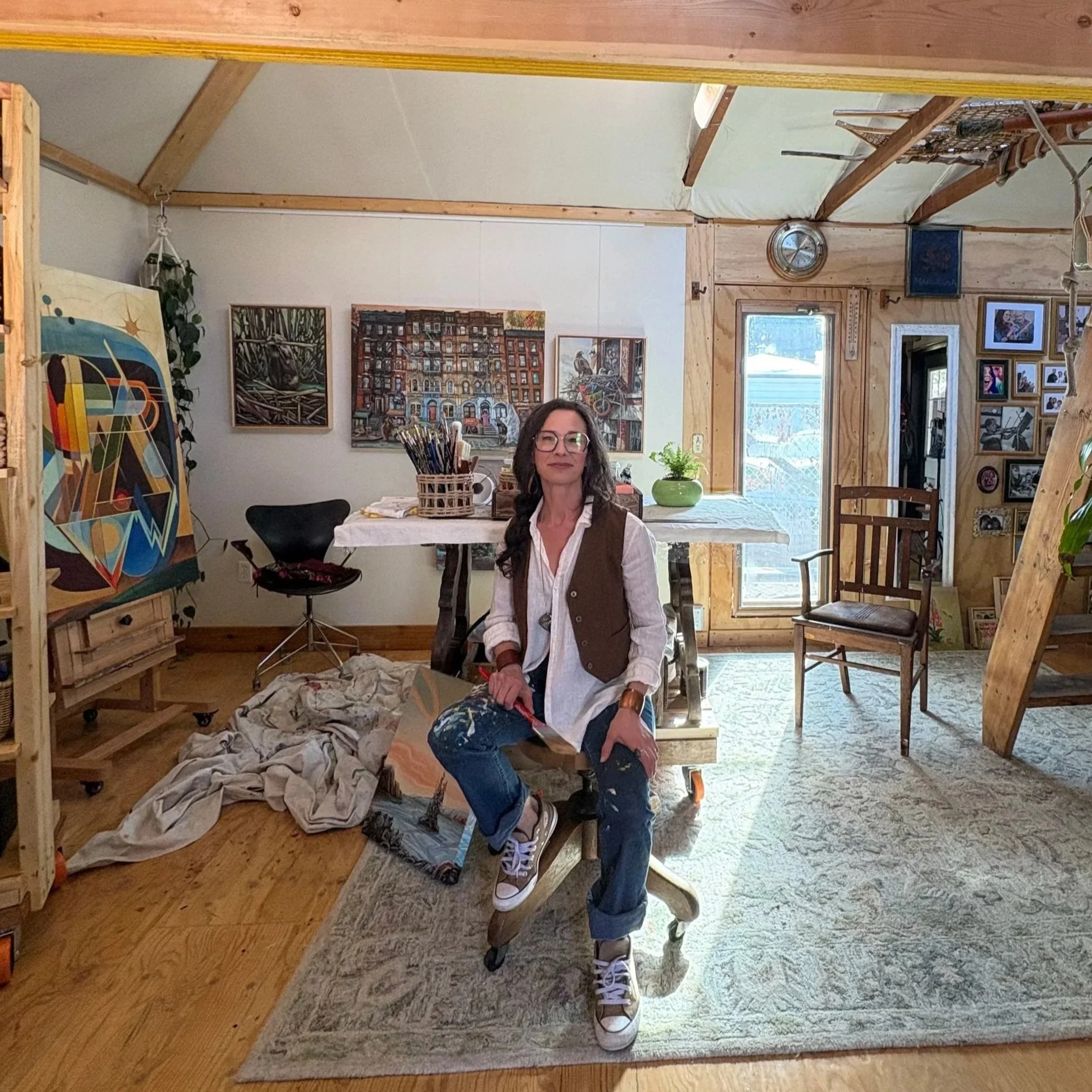 Interior of an art studio with paintings on the walls, a woman with glasses sitting at a desk, and art supplies and furniture visible, featuring wooden framing, windows, and framed photographs.