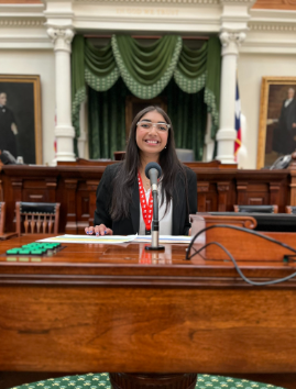 A young woman sitting at a judge's bench, smiling, in a formal government room with portraits and drapery in the background.