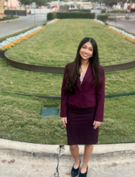 A woman in a purple blazer and black skirt standing on a sidewalk in front of a grassy park with a circular flower bed border.