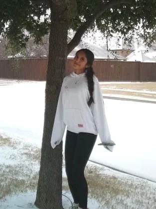 A woman with long dark hair in a ponytail standing outside next to a tree in a snowy landscape, wearing a white jacket and dark pants.
