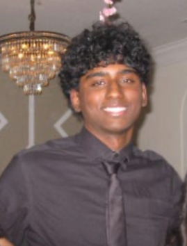 A young man with curly hair smiling, dressed in a black shirt and tie, standing indoors next to a chandelier.