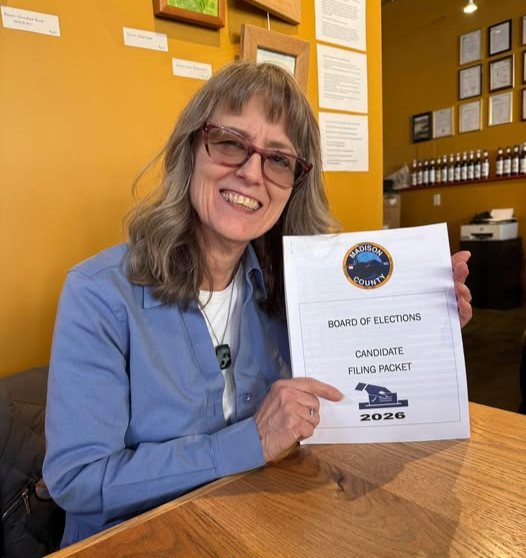 Connie Molland, Candidate for Madison County Commissioner 2026 smiling and holding a paper titled 'Madison County Board of Elections Candidate Filing Packet 2026' inside a room with yellow walls and framed documents.
