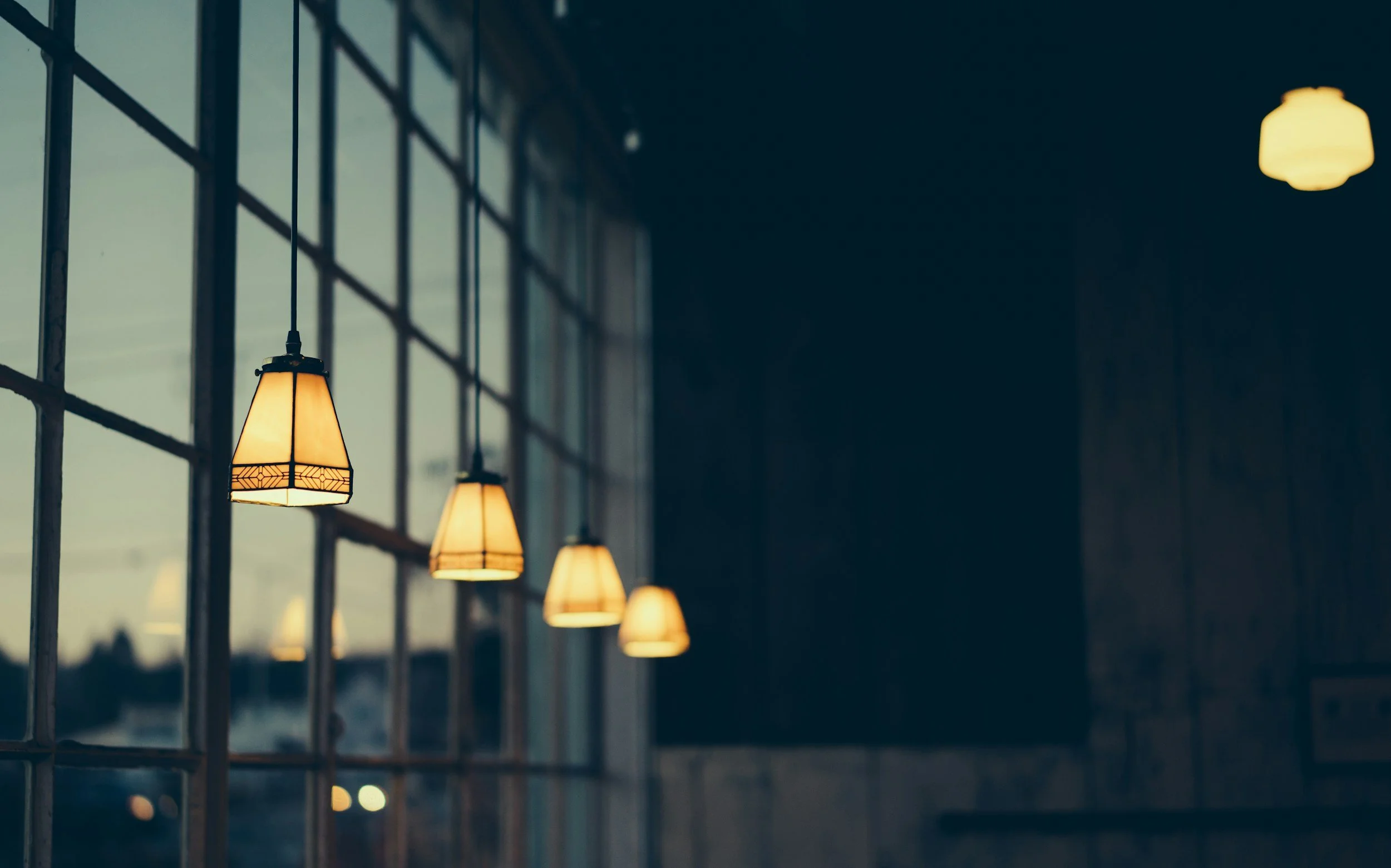 Indoor view of hanging lamps with warm light, next to a large window with grid panes, during evening or dusk.