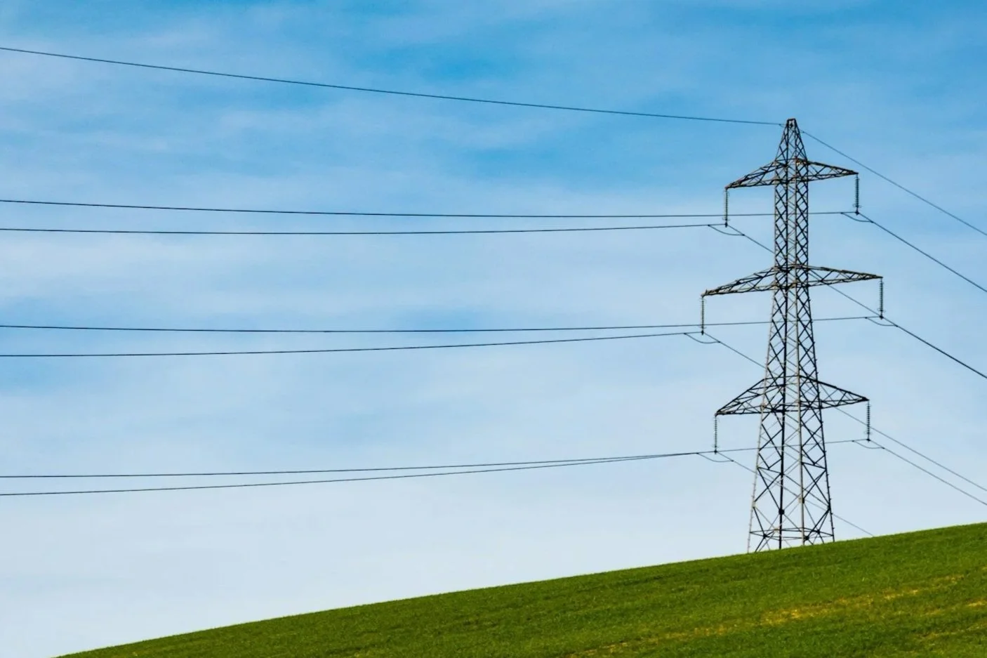 A green grassy hill with a large electrical transmission tower and power lines against a blue sky with some wispy clouds.