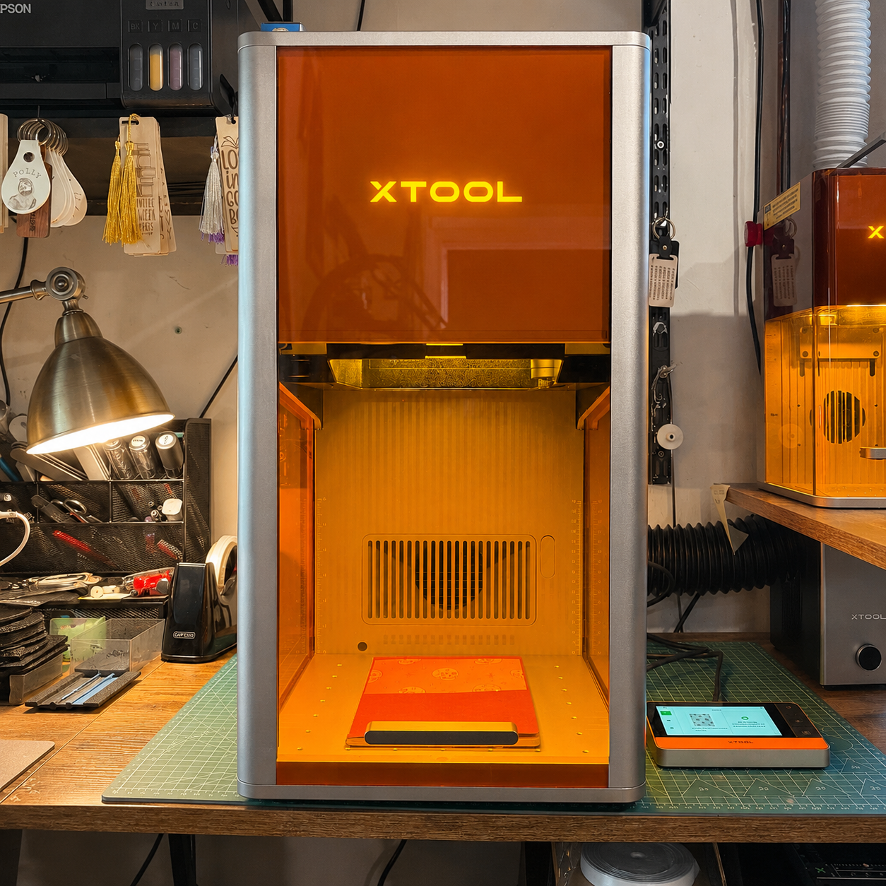 A 3D printer with an orange cover, labeled XTOOL, on a workbench in a workshop surrounded by tools and equipment.
