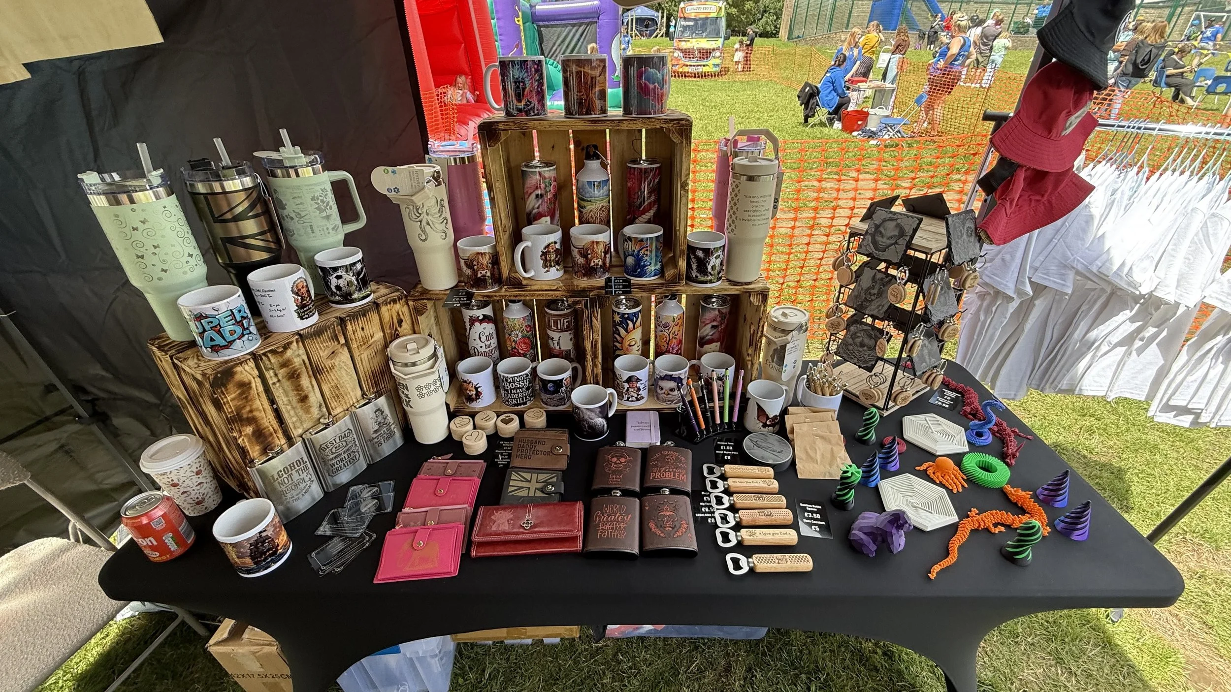 Outdoor market stall with ceramic mugs, tumblers, and small gifts, set up at a fair with people and a bounce house in the background.